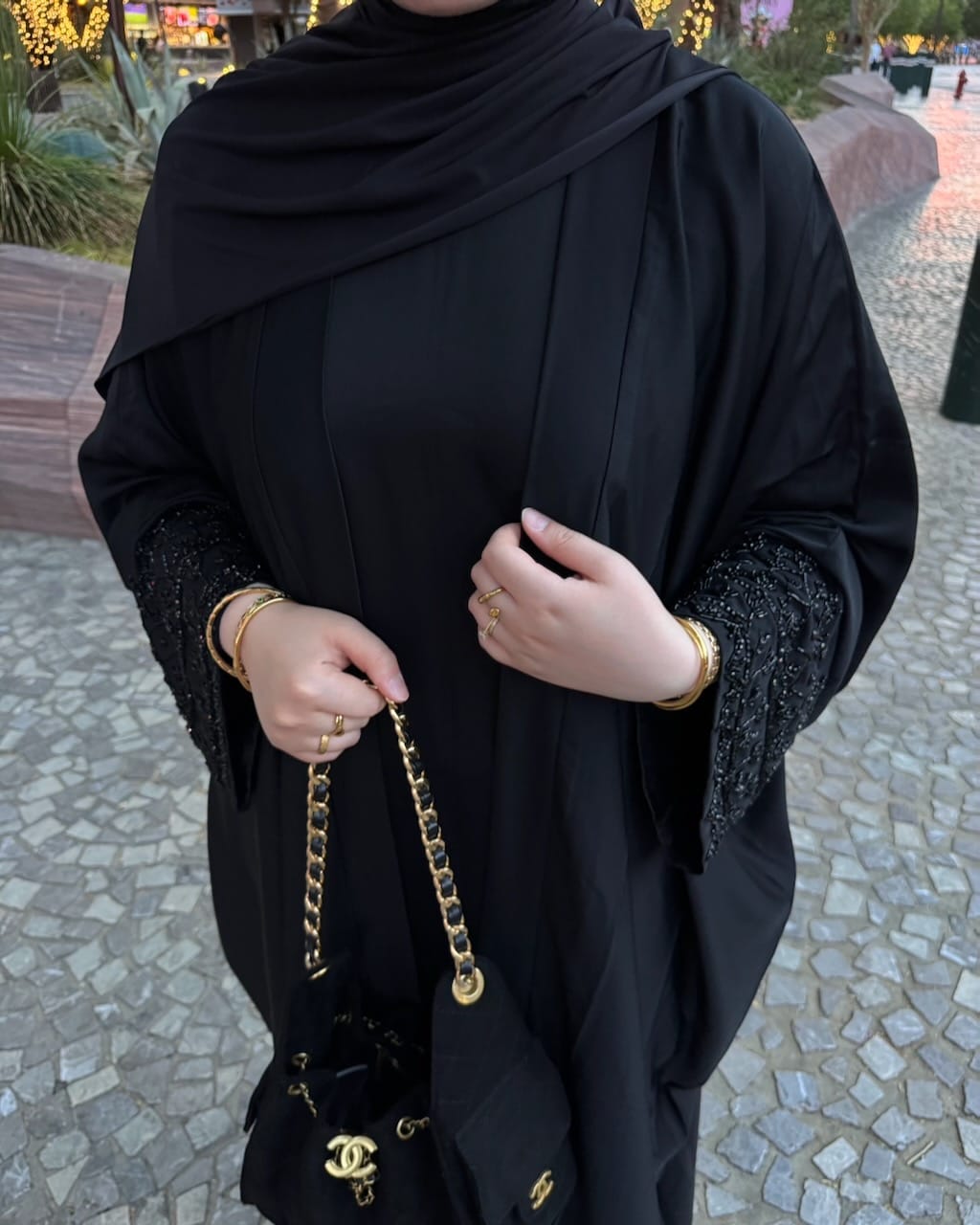 A person wearing a black abaya with textured beaded sleeves and a matching black hijab, holding a black handbag with gold chain straps in an outdoor paved setting.