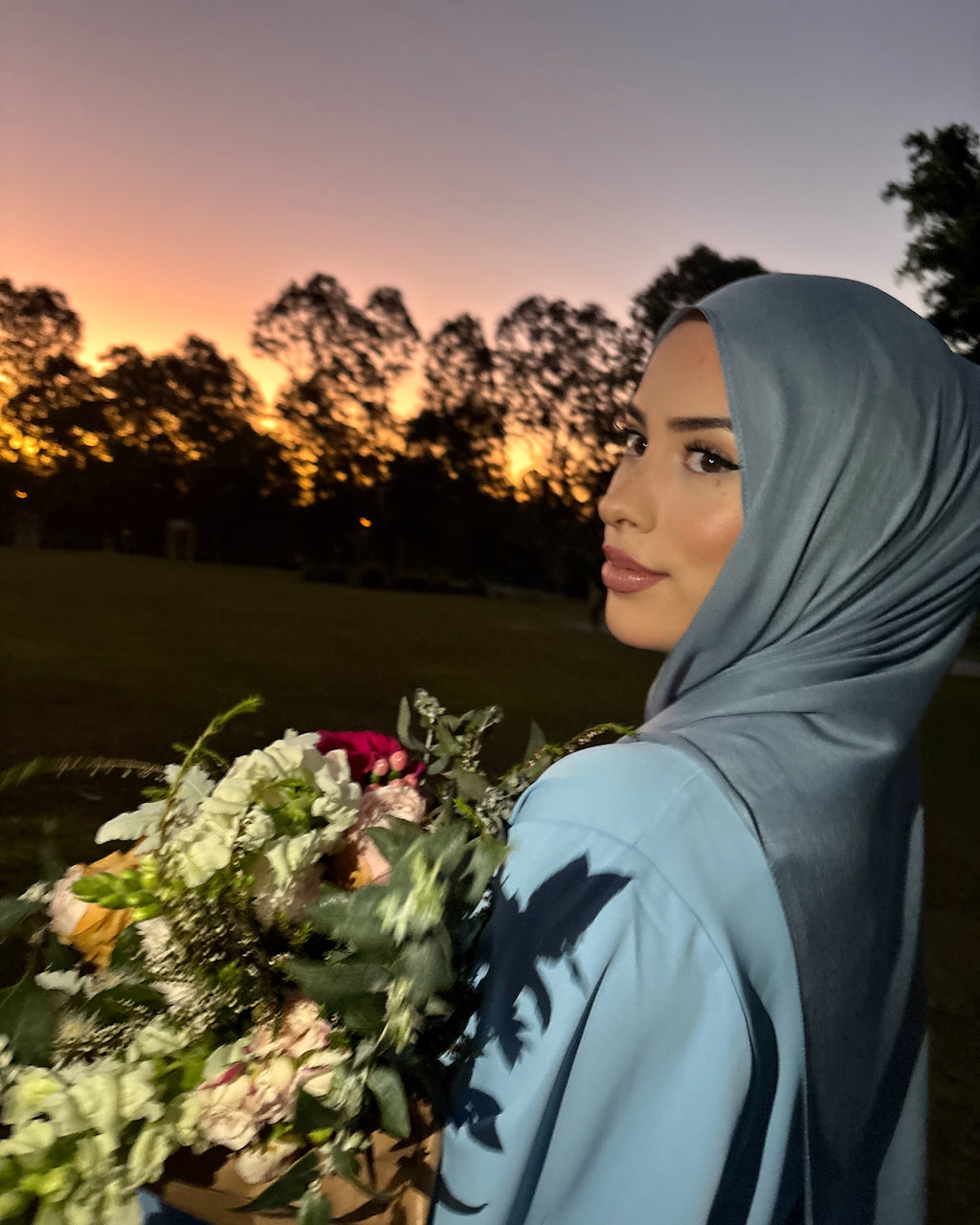 A woman wearing a light blue hijab and matching long-sleeve top, holding a large bouquet of flowers outdoors at sunset with trees in the background.