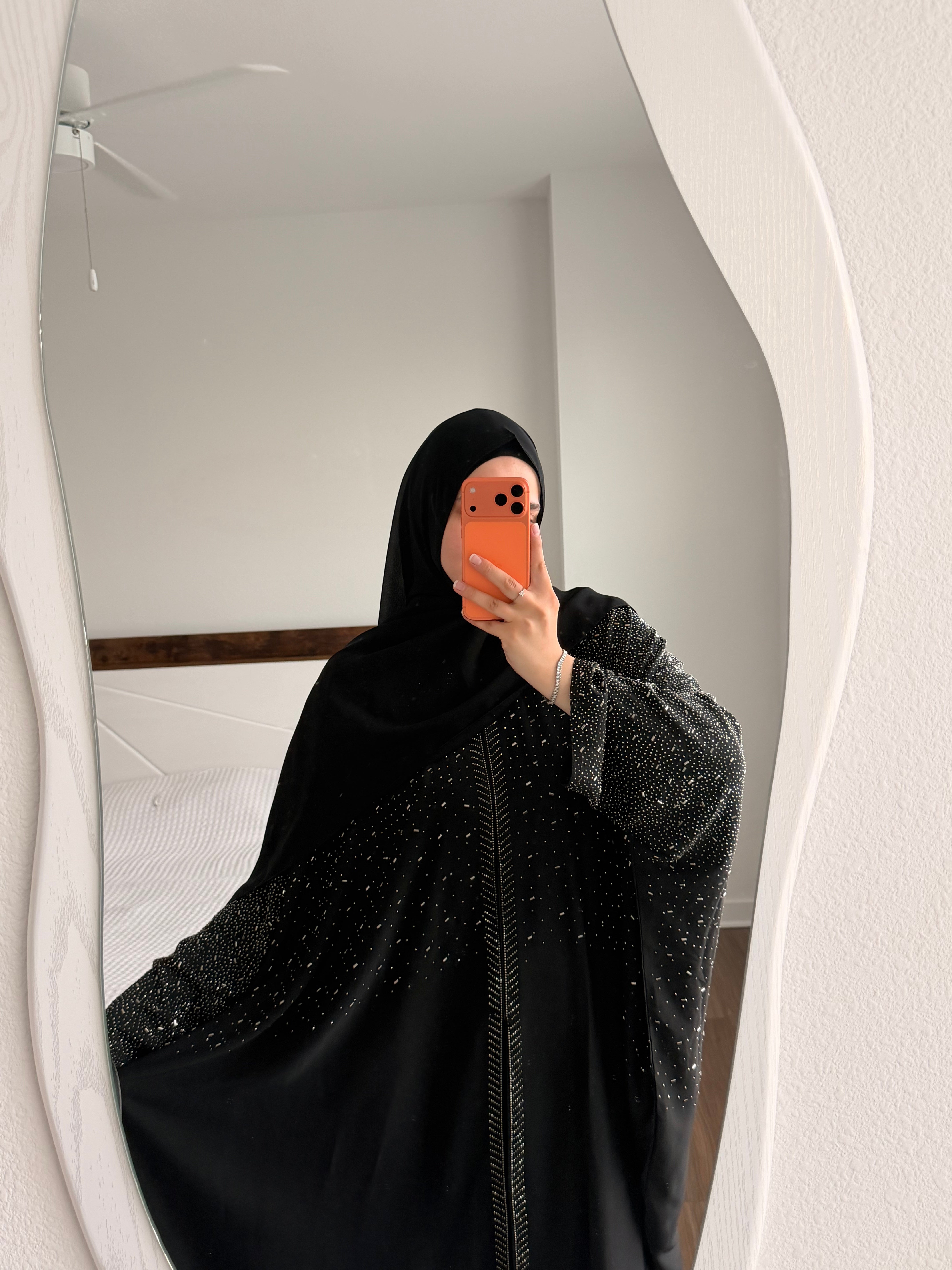 A person taking a mirror selfie in a bedroom, wearing a black full-length modest garment with a matching head covering and long sheer sleeves decorated with small silver embellishments.
