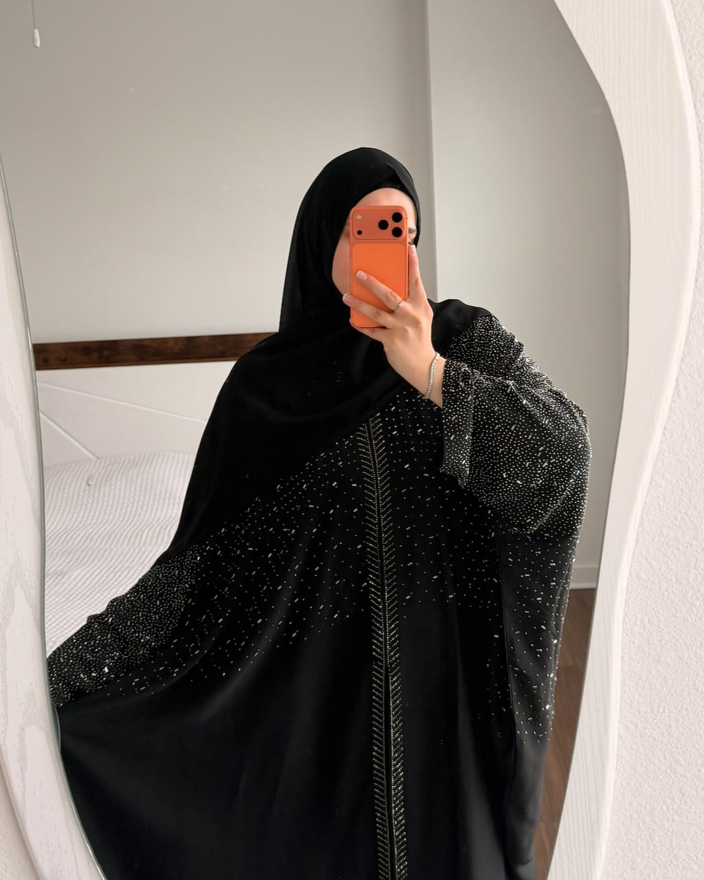 A woman takes a mirror selfie wearing a black abaya with sparkling silver embellishments and a matching black hijab. The photo is taken in a bedroom setting with a white bed and wall visible behind her.