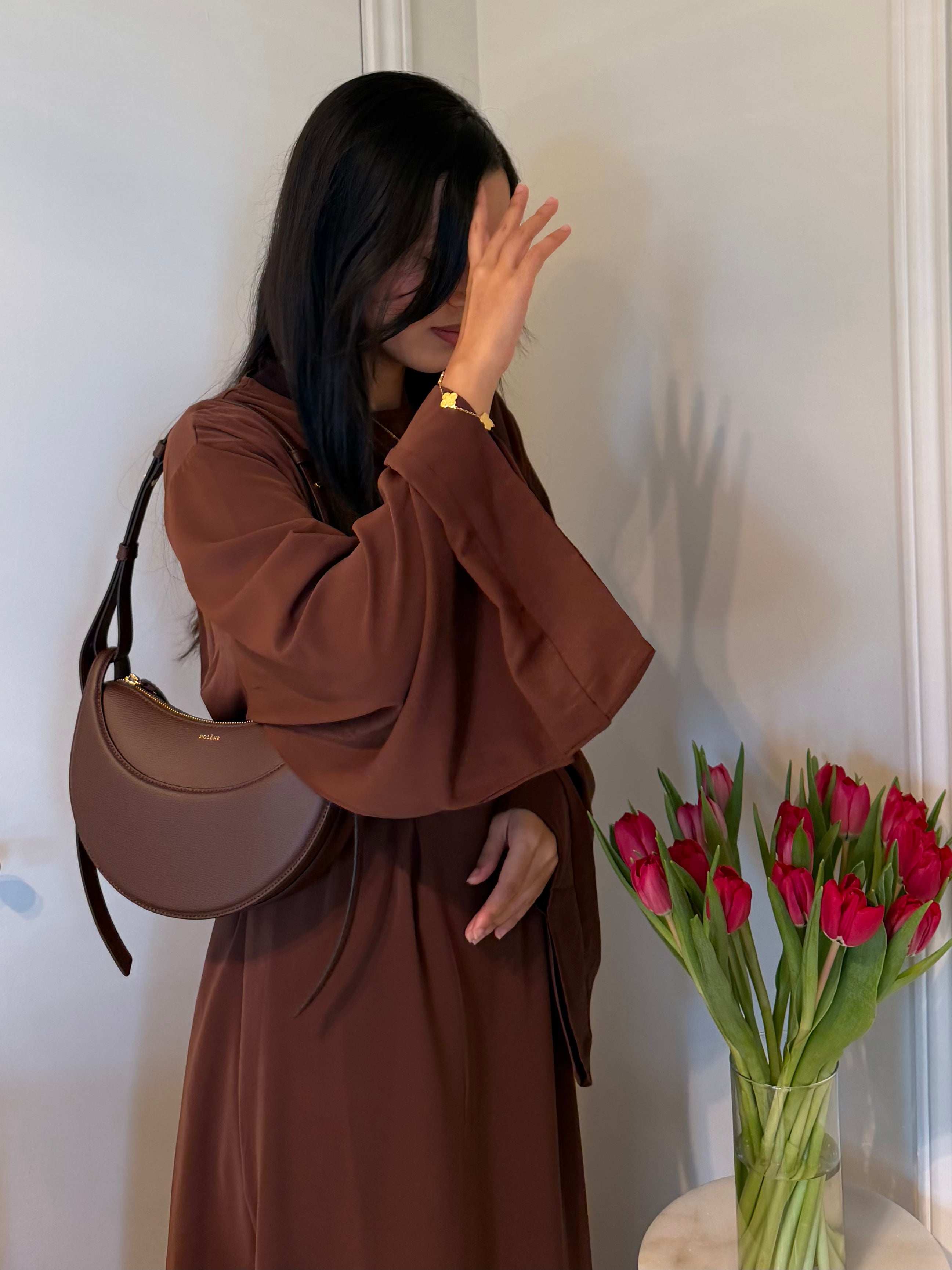 A woman wearing a long-sleeved brown dress and carrying a brown crescent-shaped shoulder bag, standing indoors next to a vase of pink tulips.