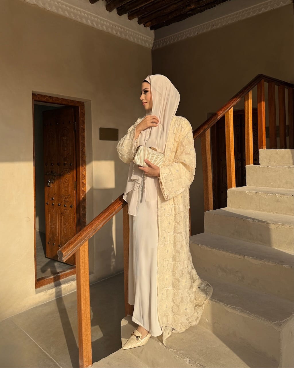A woman wearing a traditional modest outfit with a cream-colored embroidered robe, matching headscarf, and pointed flats, standing on stone stairs in a sunlit building with wooden railings.