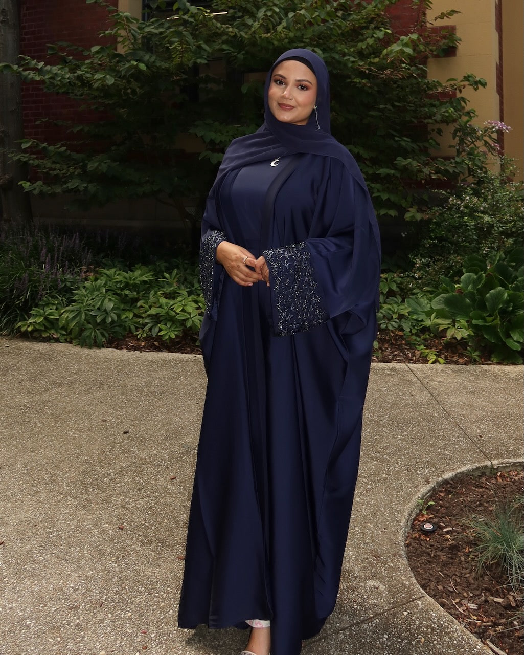 A woman wearing a navy blue modest outfit with a matching headscarf and a long abaya featuring decorative silver embroidery on the sleeves, standing outdoors on a paved walkway with green plants and a building in the background.