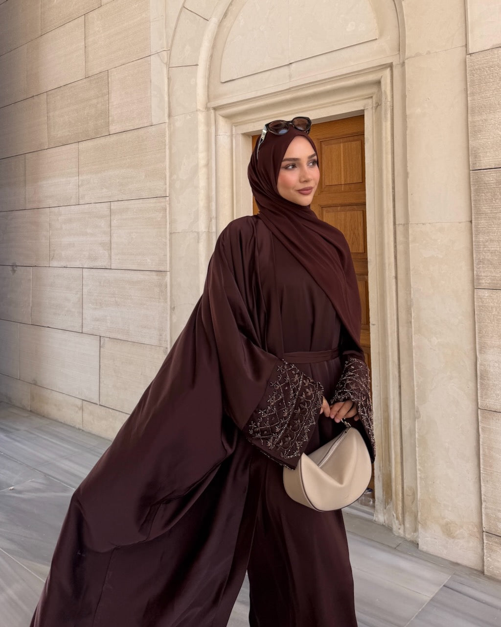 A woman wearing a dark brown modest outfit with a matching hijab and sunglasses on her head, holding a beige crescent-shaped handbag, standing in front of a beige stone archway.