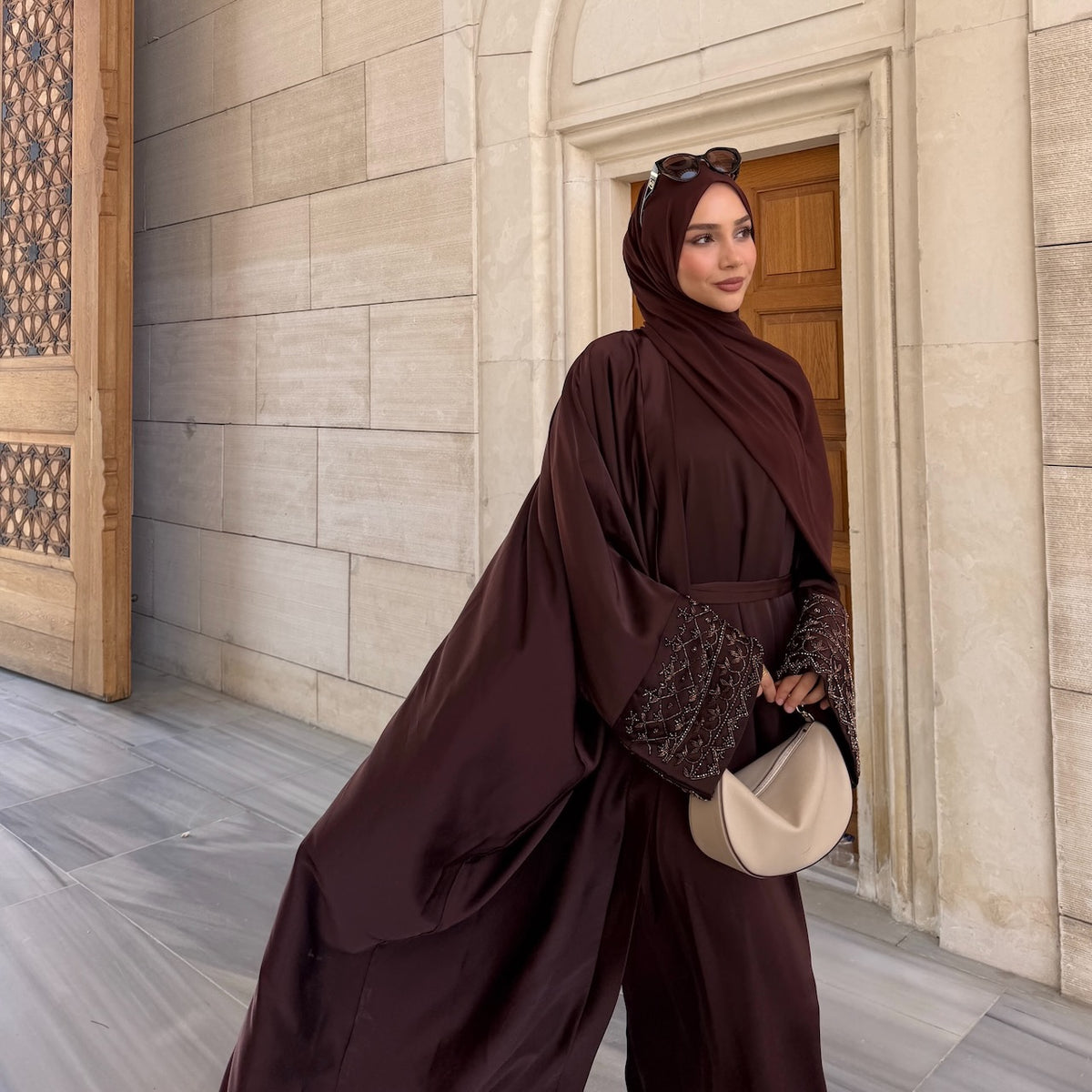 a women wearing modest clothing, a coffee coloured abaya with handbag in outdoor stone courtyard settings. Wearing sunglases and makeup. going to eid.