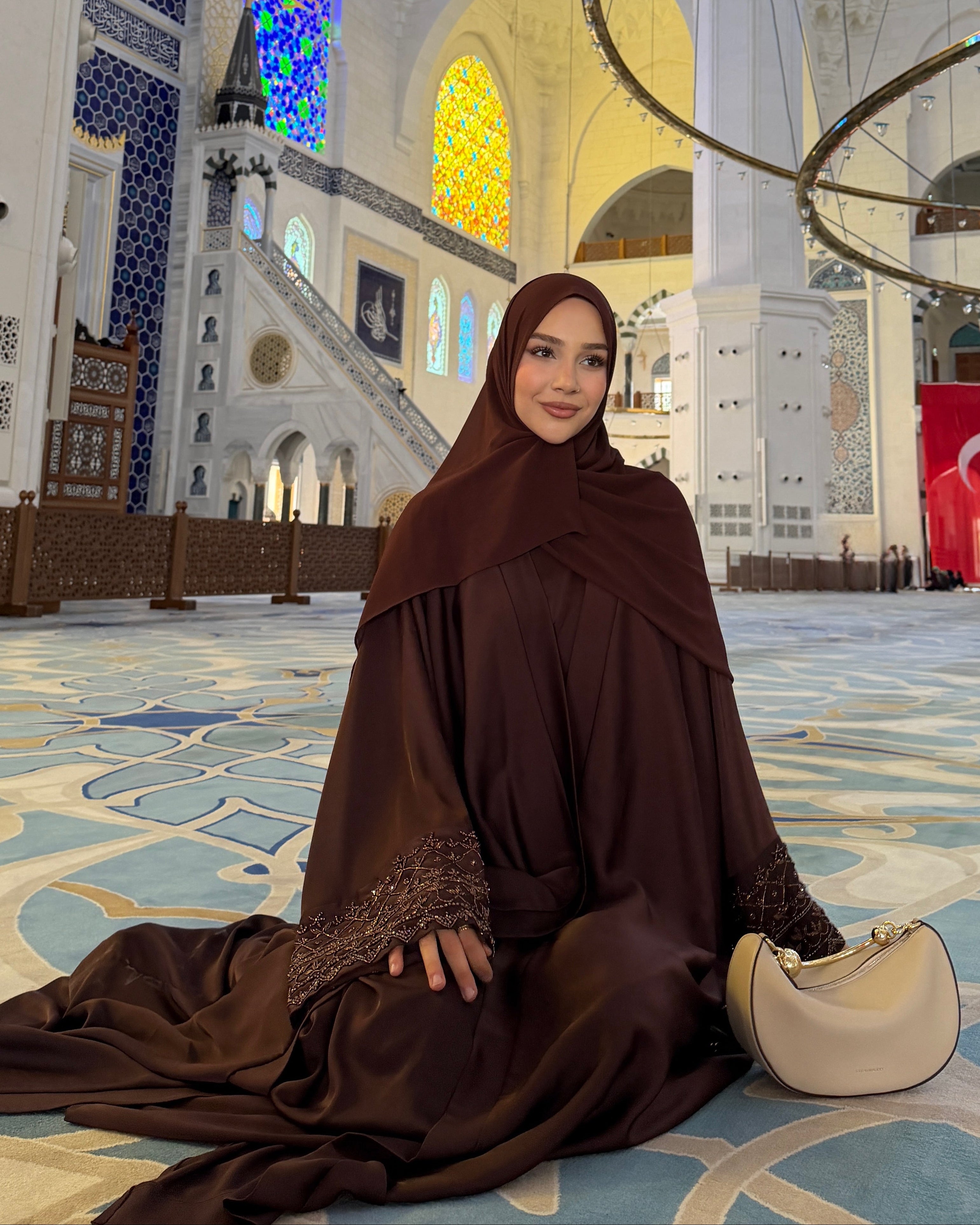 Woman in a brown dress sitting inside a grand mosque with intricate architecture.