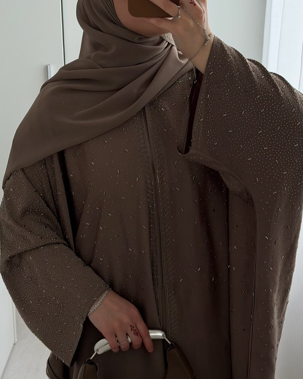 A person wearing a brown abaya with subtle silver speckled details and a matching brown hijab, holding a brown handbag in a bright indoor setting.