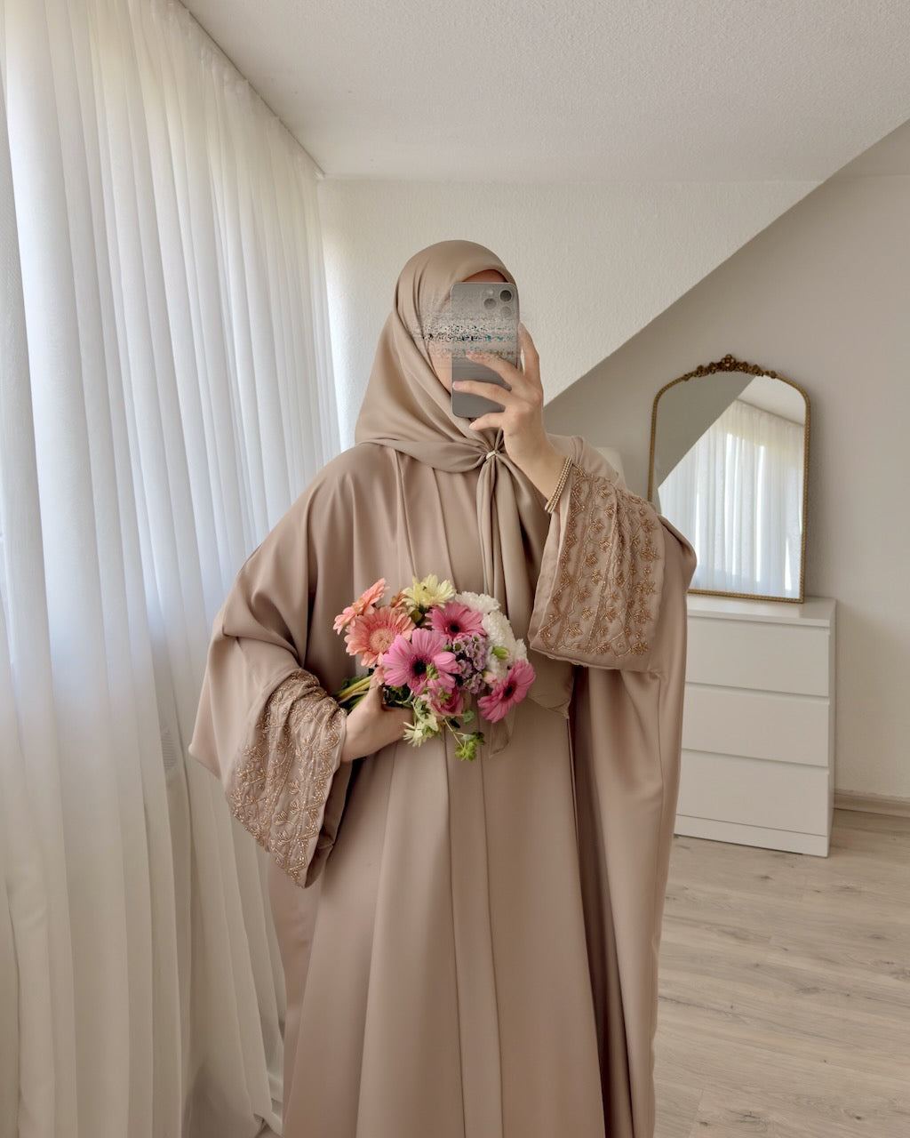 A woman wearing a beige modest outfit with a matching hijab and long lace-trimmed sleeves, taking a mirror selfie in a bright room with white curtains, a dresser, and a mirror. She is holding a bouquet of pink and white flowers.