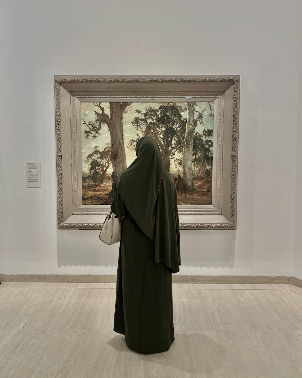 A person wearing a dark olive green abaya and khimar stands in a gallery, facing a large framed landscape painting of trees.