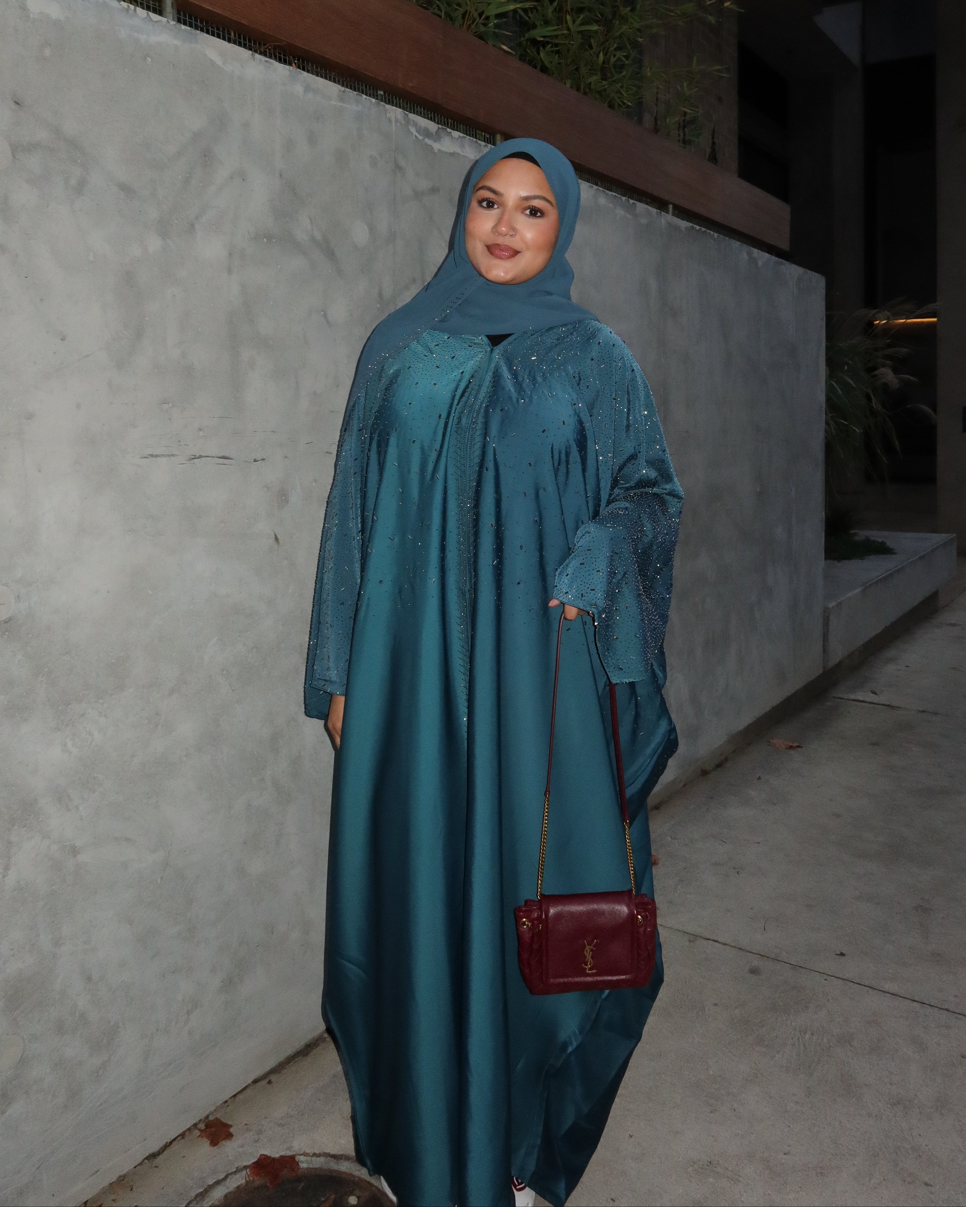 A woman wearing a teal blue abaya and matching hijab stands outdoors at night next to a concrete wall, holding a small burgundy handbag.