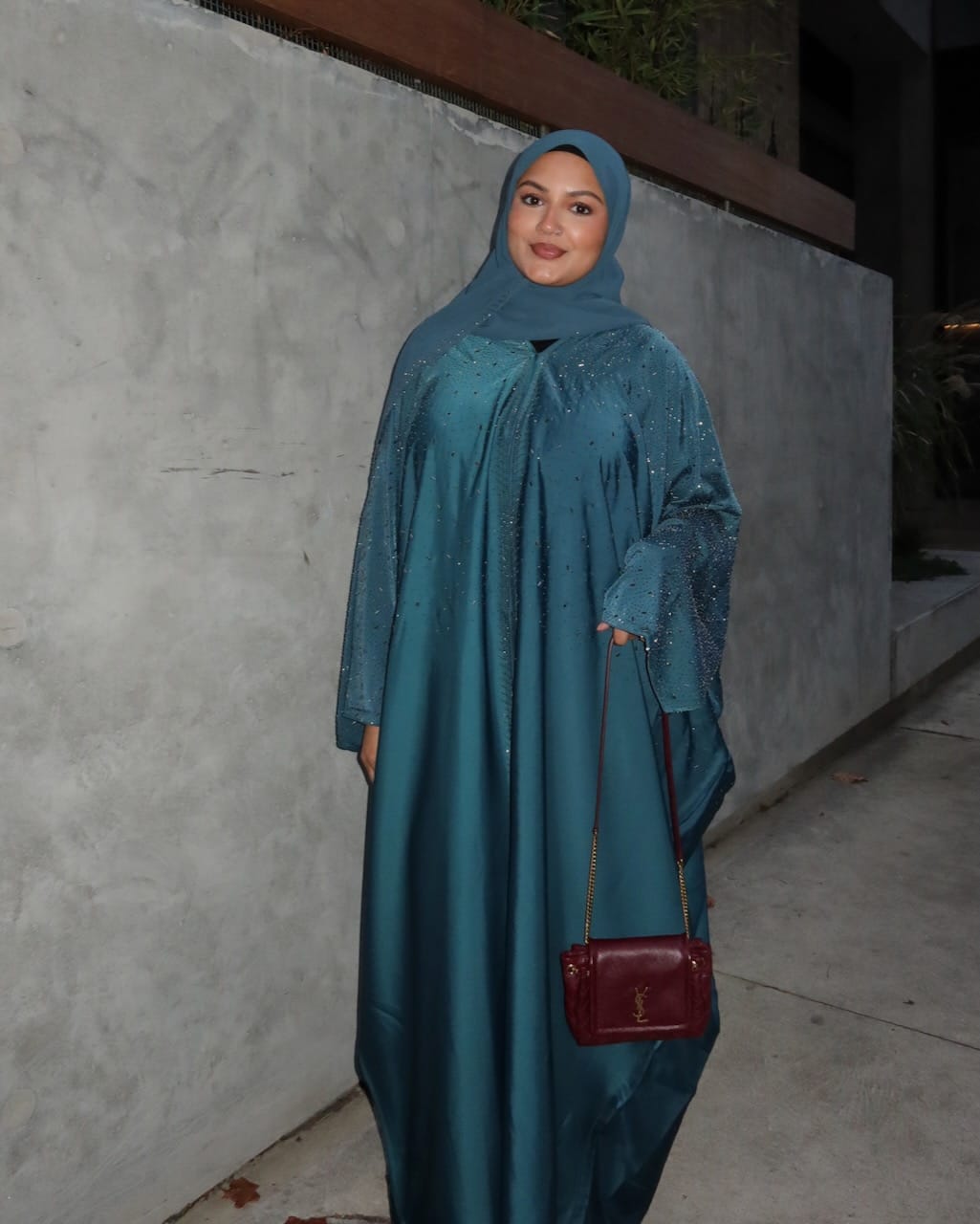 A woman wearing a teal blue abaya and matching hijab, holding a small burgundy shoulder bag, standing against a textured gray wall at night.