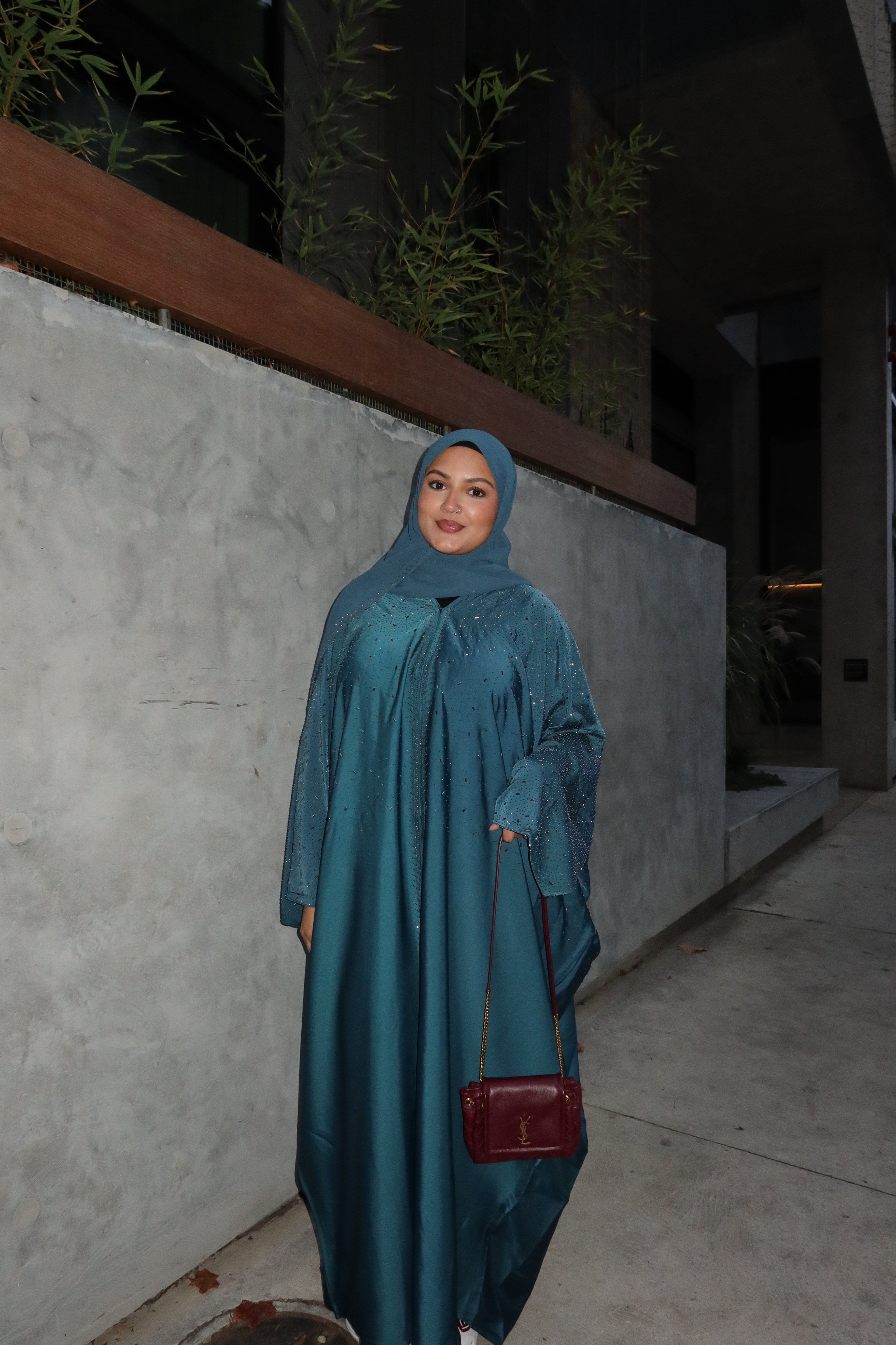 A woman wearing a teal blue abaya and matching hijab stands outdoors at night next to a concrete wall, holding a small burgundy handbag.