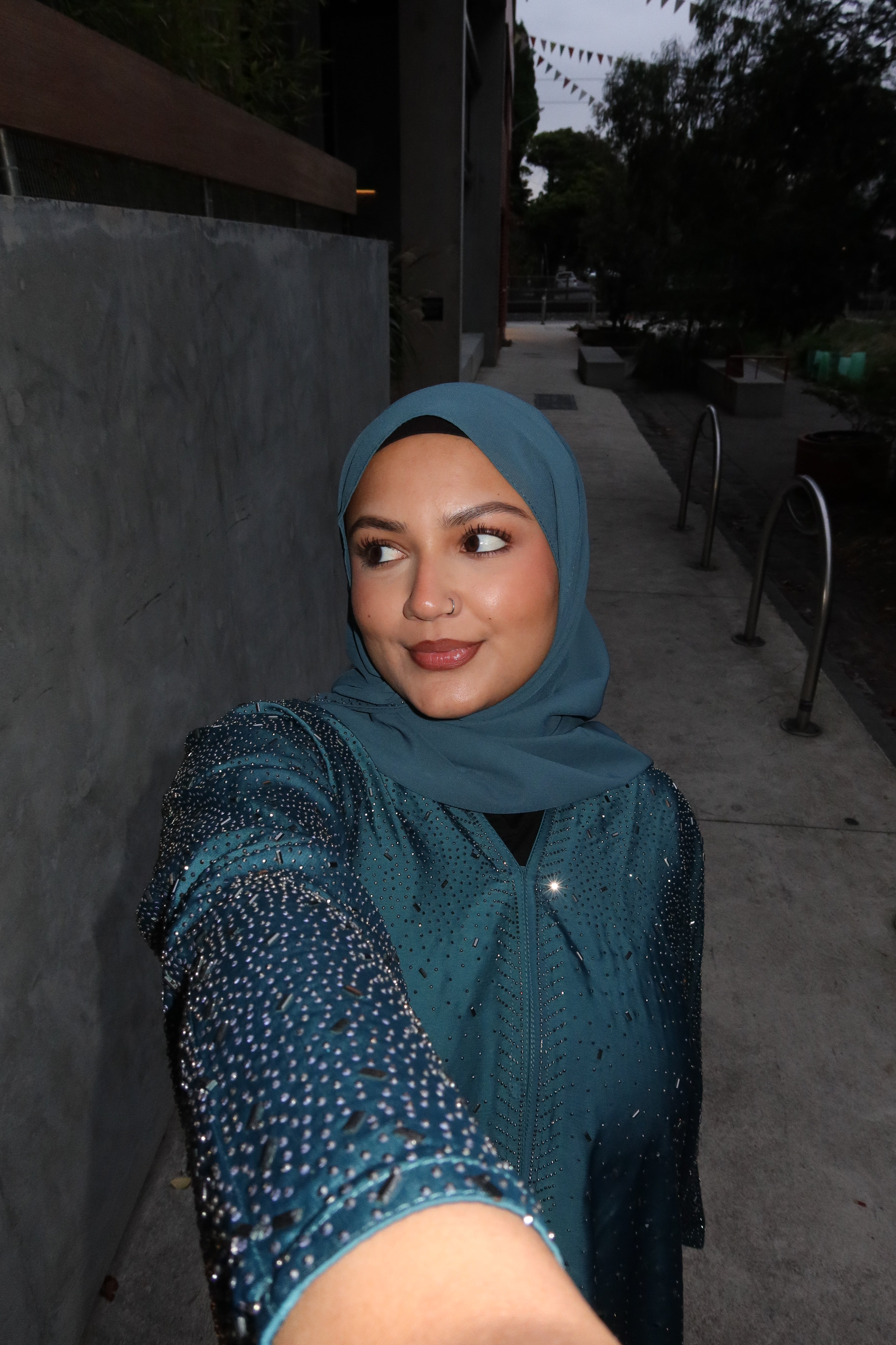 A woman wearing a teal blue hijab and a matching embellished teal top takes a selfie in an outdoor alleyway at dusk.