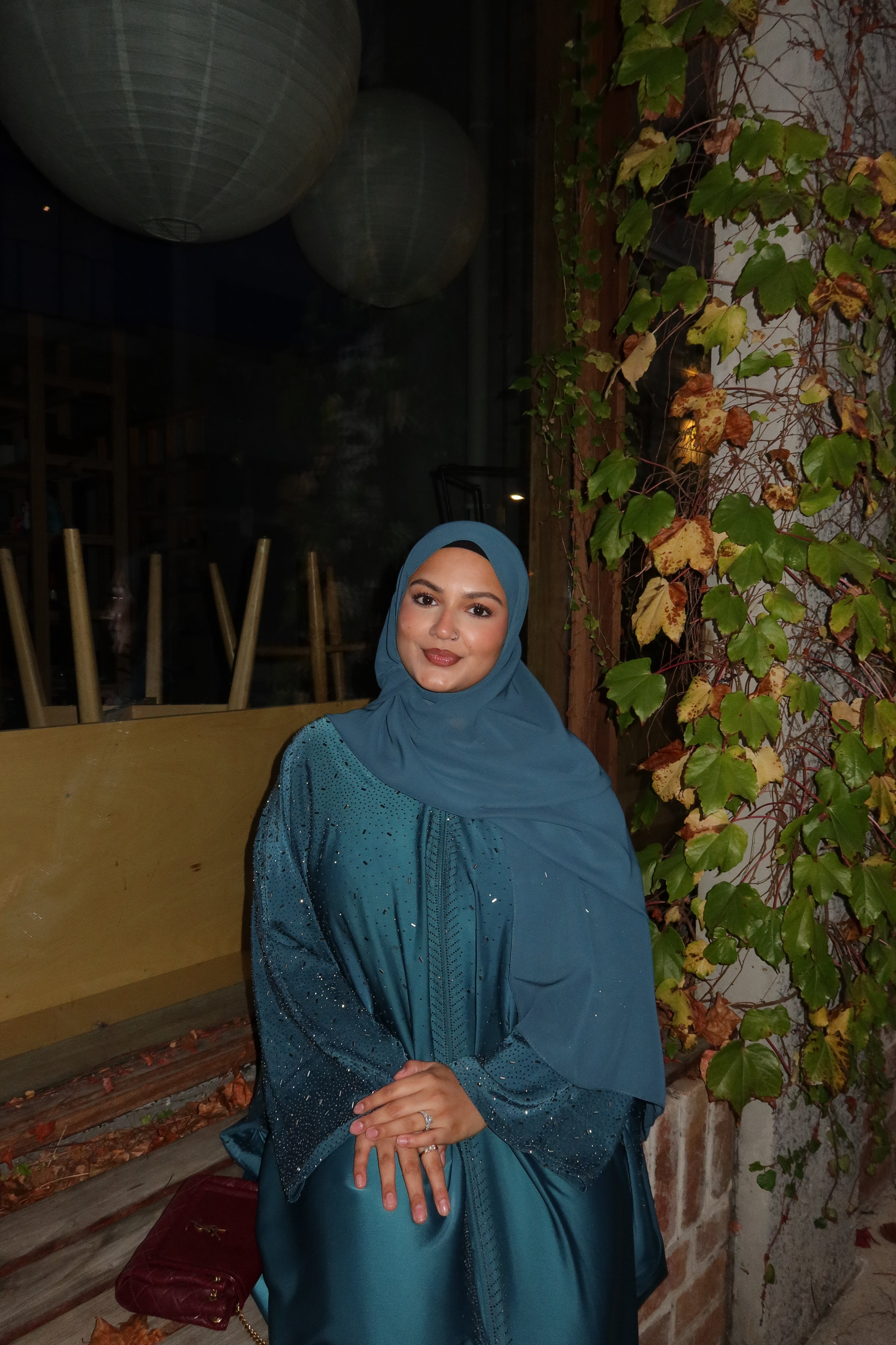 A woman wearing a teal blue abaya and matching hijab stands outdoors at night in front of a wall with ivy and hanging paper lanterns.