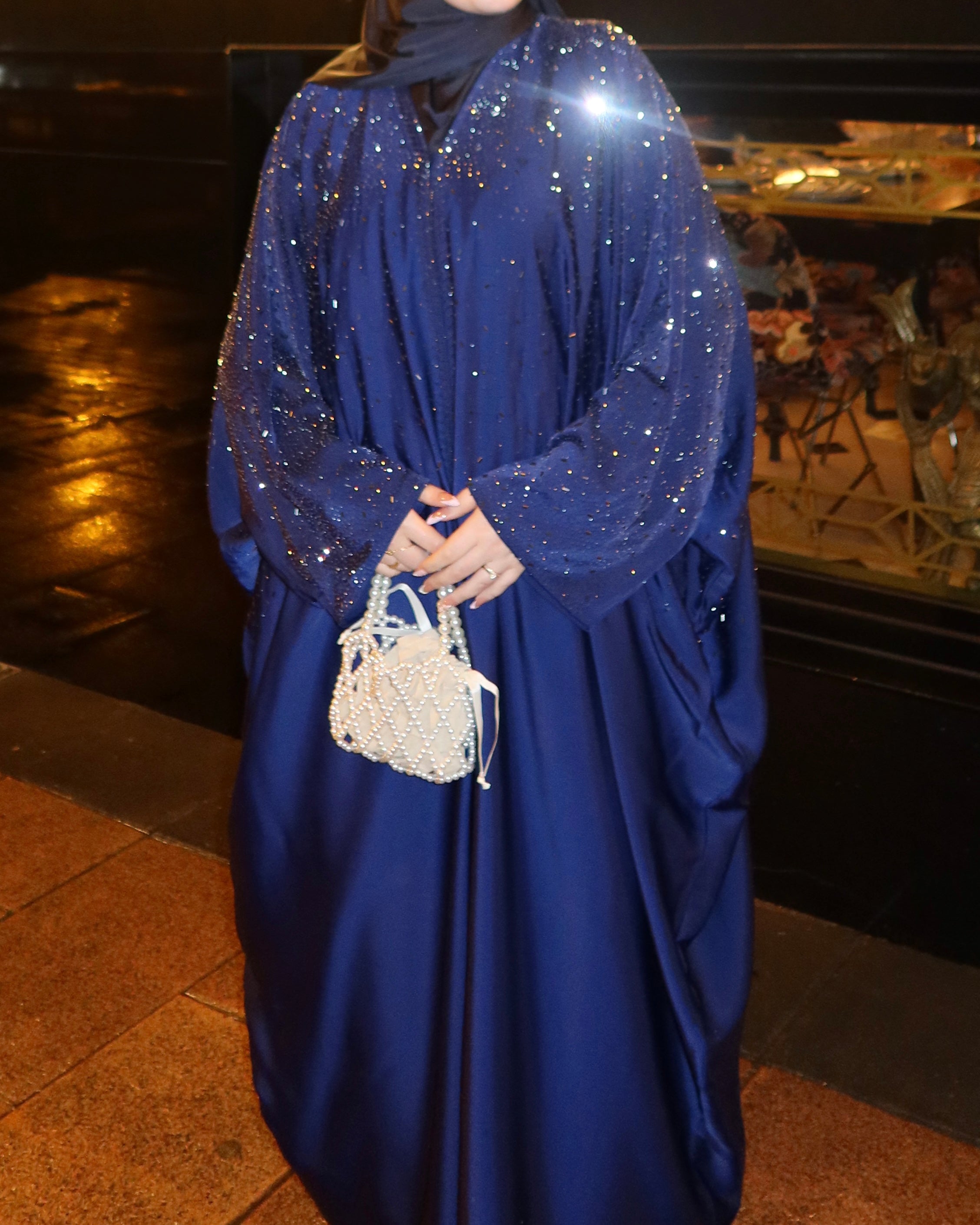 A person wearing a deep blue abaya with sparkling sequined sleeves and a matching headscarf, holding a small white beaded handbag in an outdoor nighttime setting.