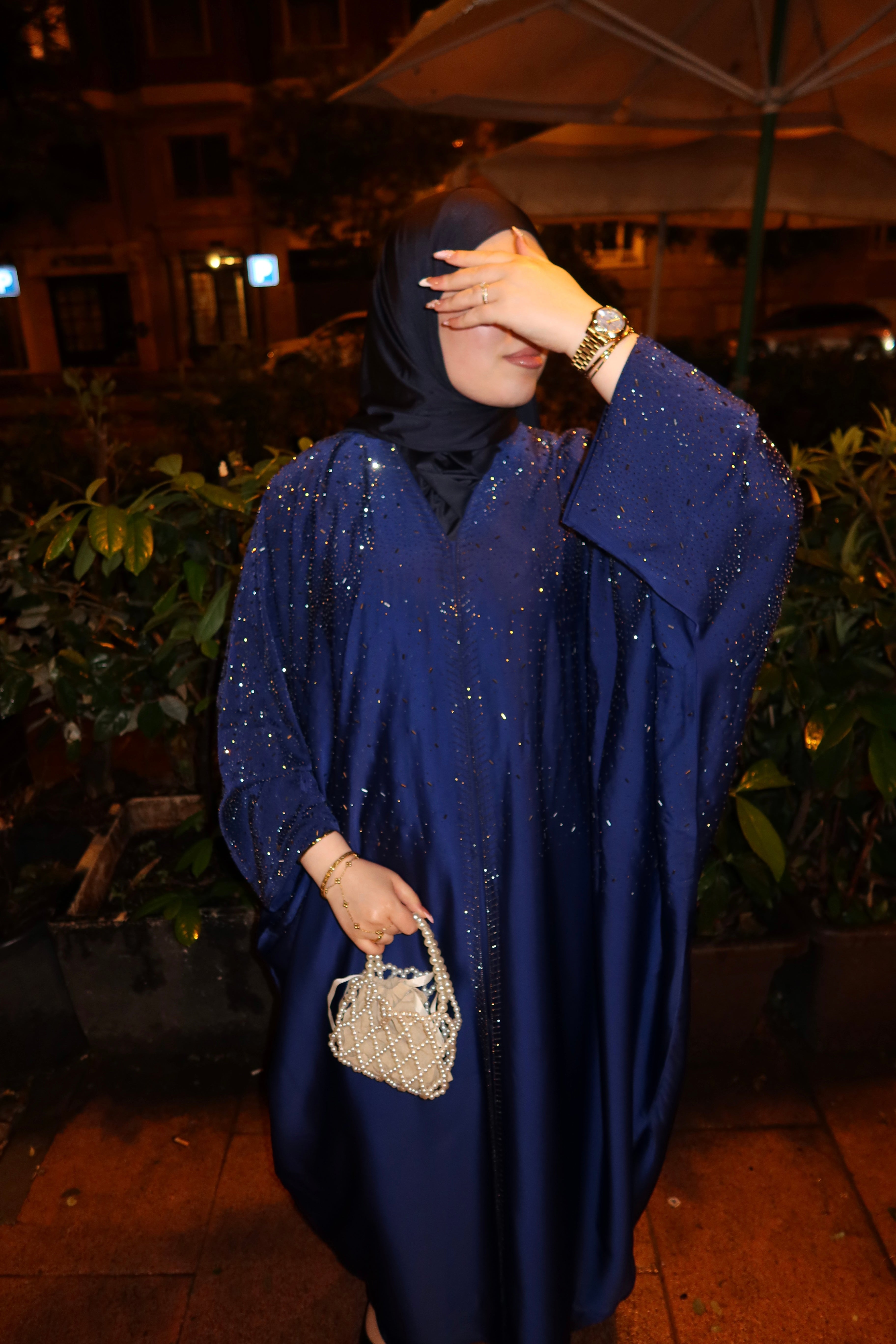 A woman wearing a sparkling royal blue abaya and black hijab stands outdoors at night, holding a small woven handbag.