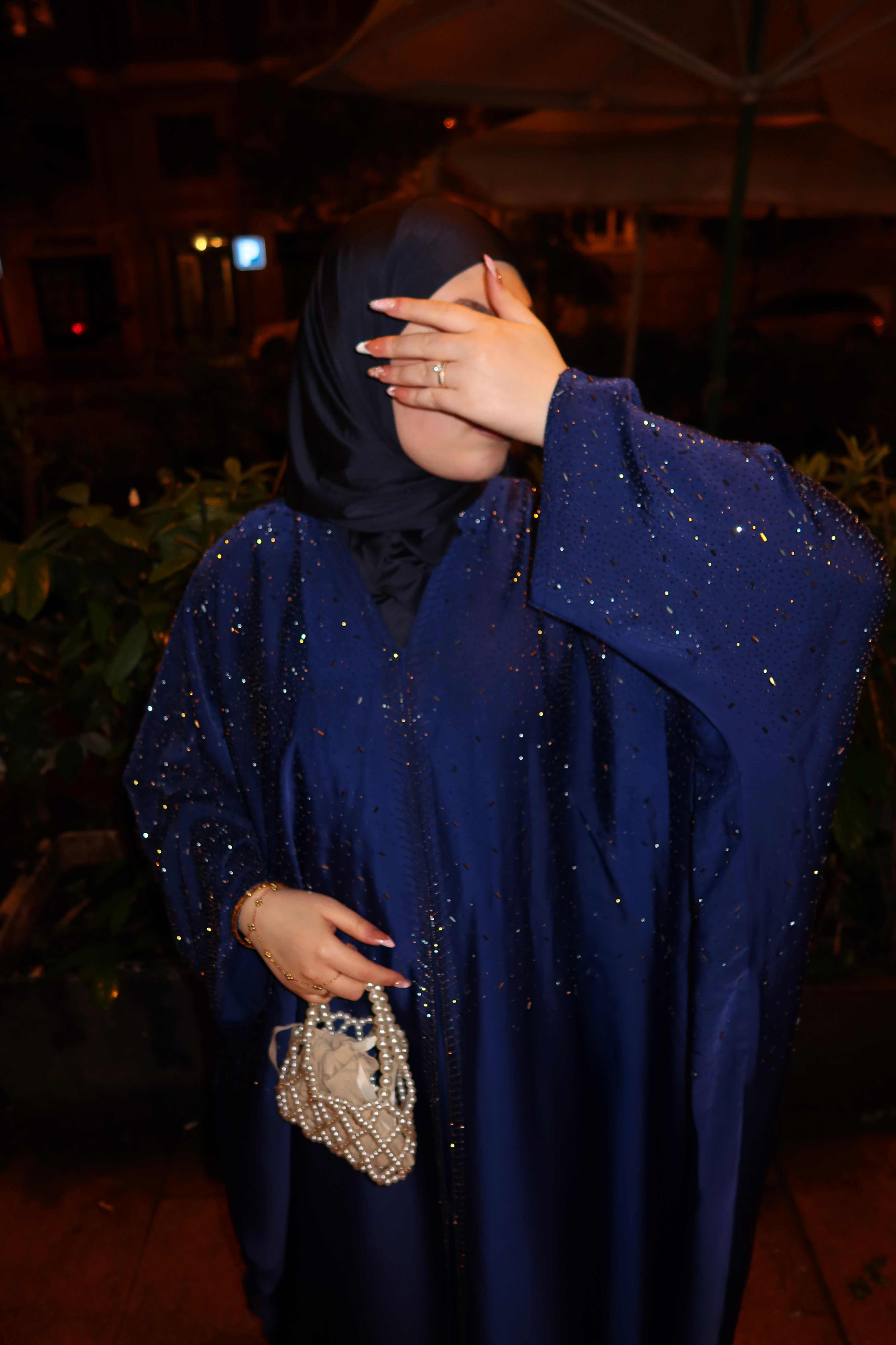 A woman wearing a sparkling royal blue abaya with a black hijab, posing at night with a small pearl-beaded handbag.