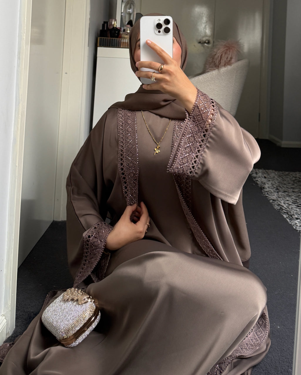 A woman takes a mirror selfie wearing a brown satin abaya with matching lace-trimmed sleeves and a coordinating headscarf. She is holding a white smartphone and carrying a small glittery clutch bag in a home setting. - Fajr Noor