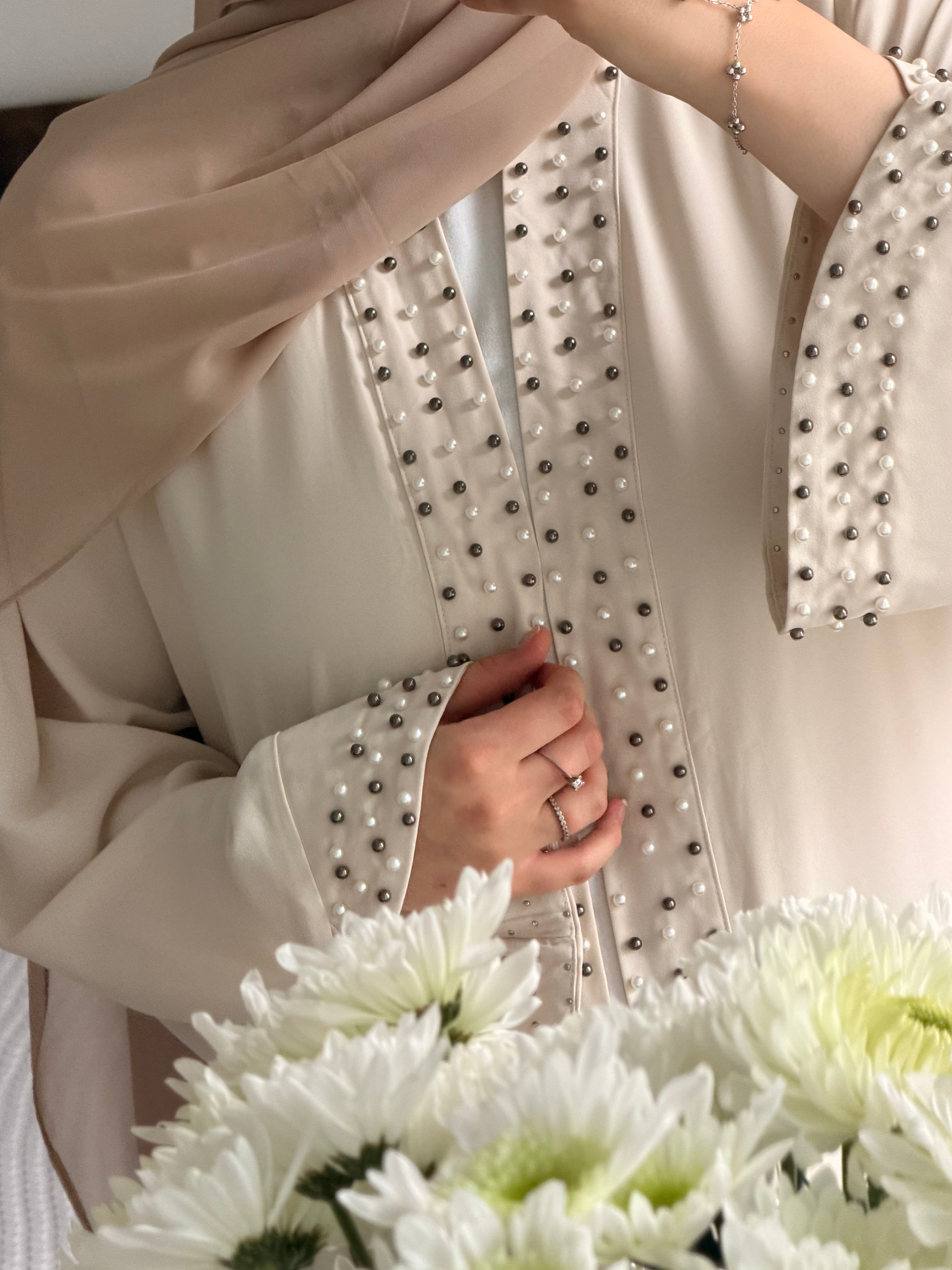 A close-up of a person wearing a beige long-sleeve abaya or tunic with pearl and metallic bead embellishments along the front and cuffs, paired with a matching beige hijab. White flowers are visible in the foreground.