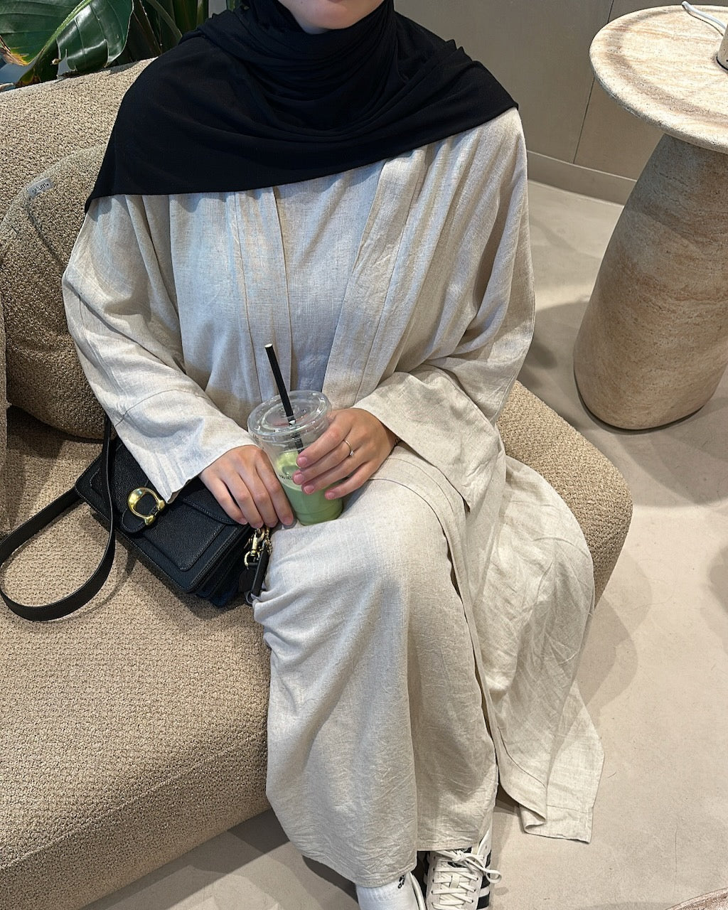 A person seated on a beige upholstered chair wearing a loose, long-sleeve abaya in a light cream or off-white tone with a black hijab. The person is holding a clear plastic drink cup with a green beverage and black straw, and a black handbag rests beside them.