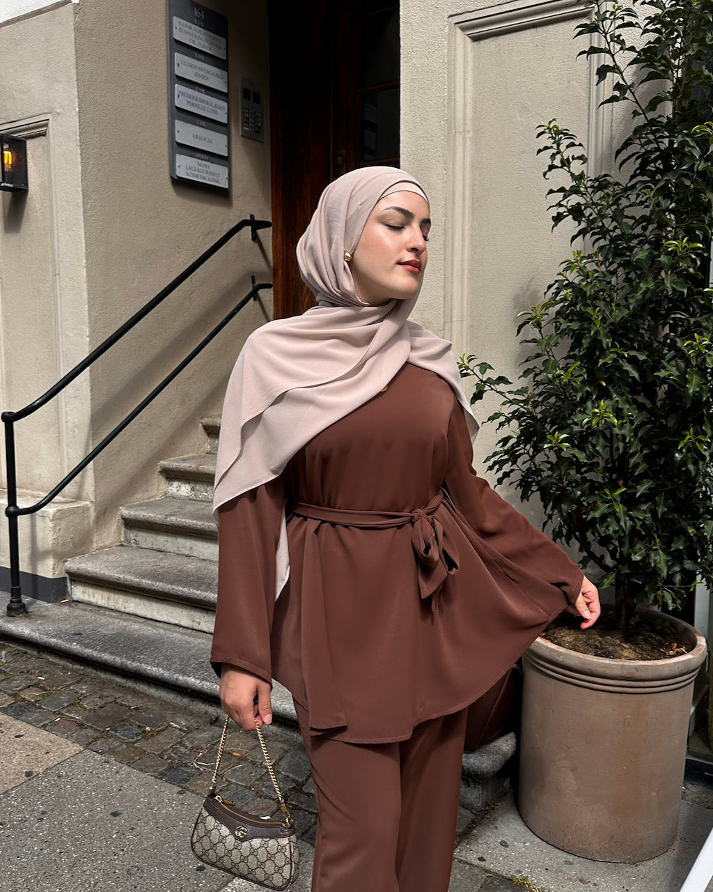 A woman wearing a matching brown two-piece outfit with a tie waist and a light beige hijab, standing outdoors in front of a building entrance with steps and a potted plant.