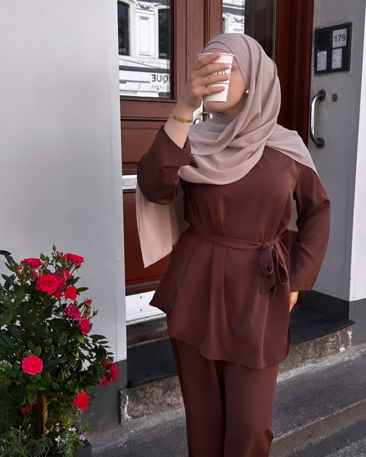 Person wearing a brown outfit and hijab taking a selfie outside a building with flowers in the foreground.