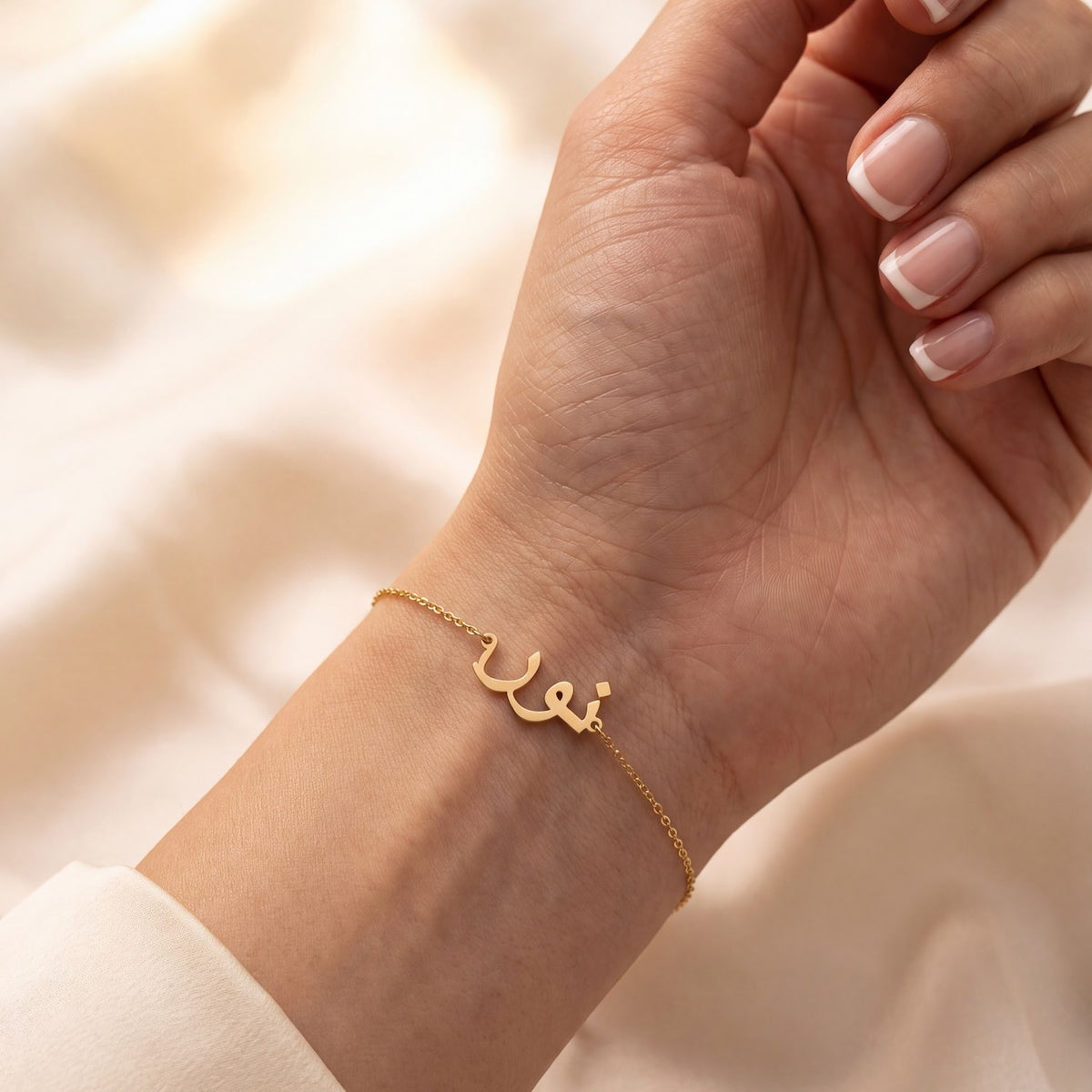 A delicate gold chain bracelet with a small Arabic calligraphy nameplate worn on a woman's wrist, shown against a soft beige fabric background.