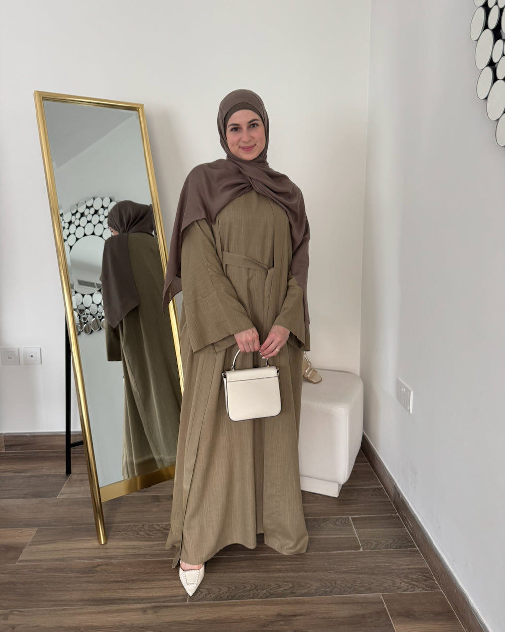 A woman wearing a taupe-brown hijab and a matching olive-brown abaya with wide sleeves, standing in a room with a gold-framed mirror, white wall, and wood-look floor. She is holding a small white handbag.