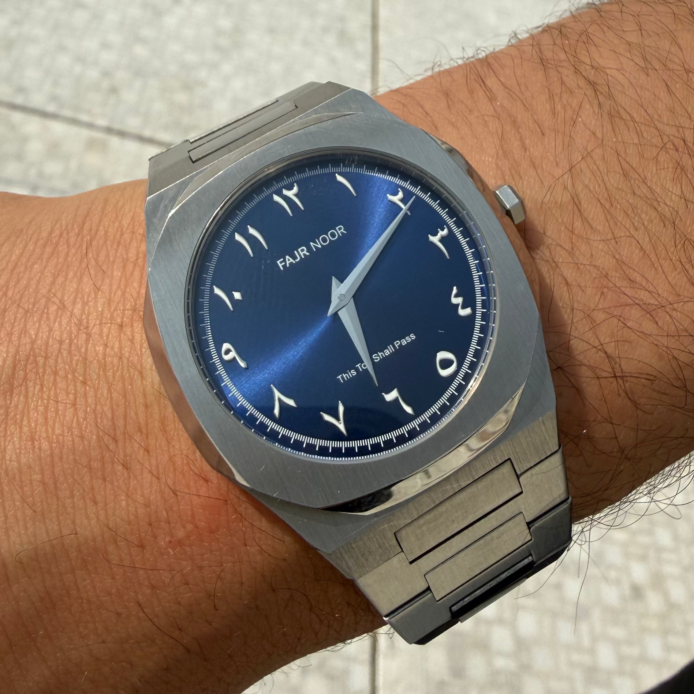 Silver watch with a blue dial on a wrist, brand 'FAJR NOOR' visible