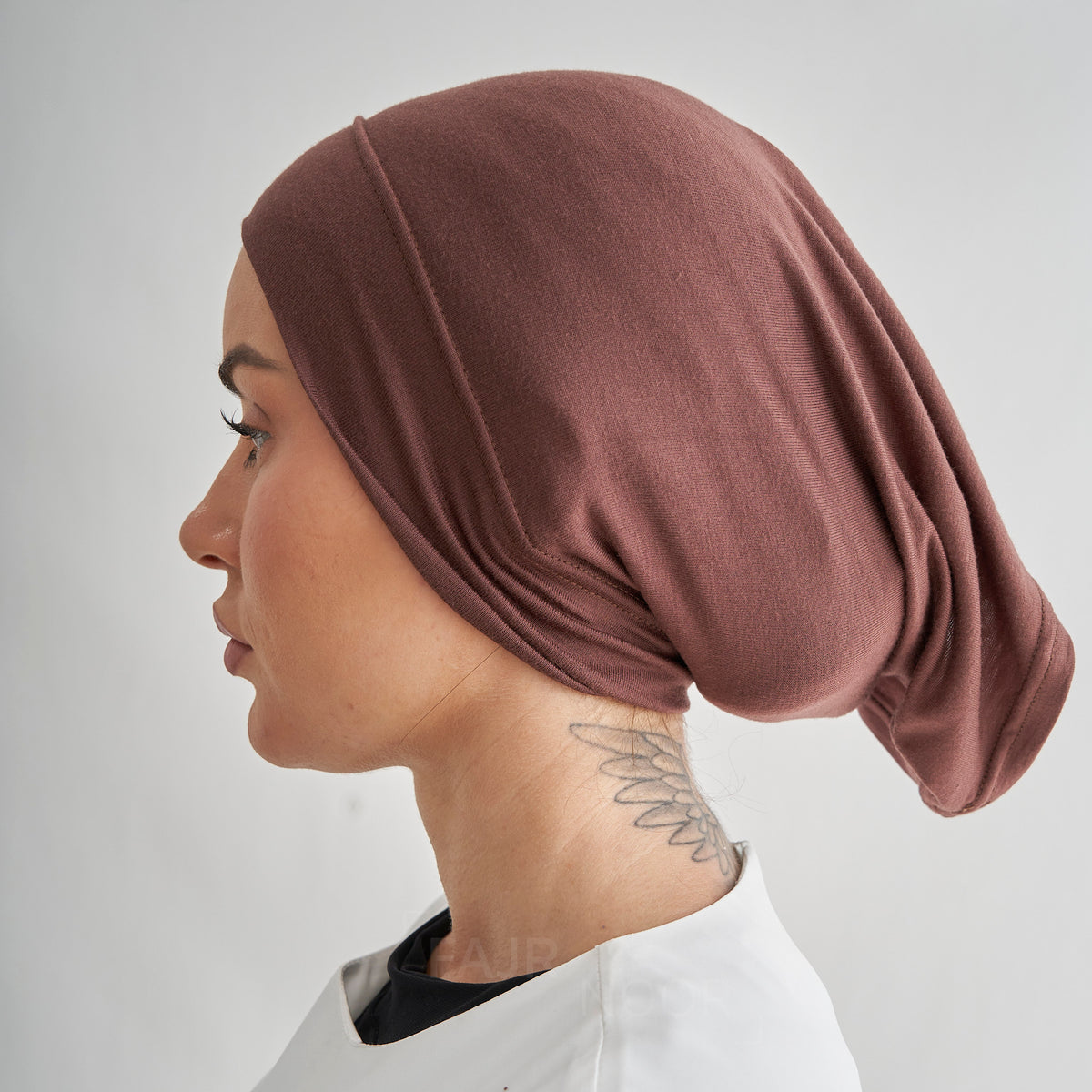 A side-profile view of a person wearing a draped, dusty rose-brown fabric head covering or hijab against a light gray background.