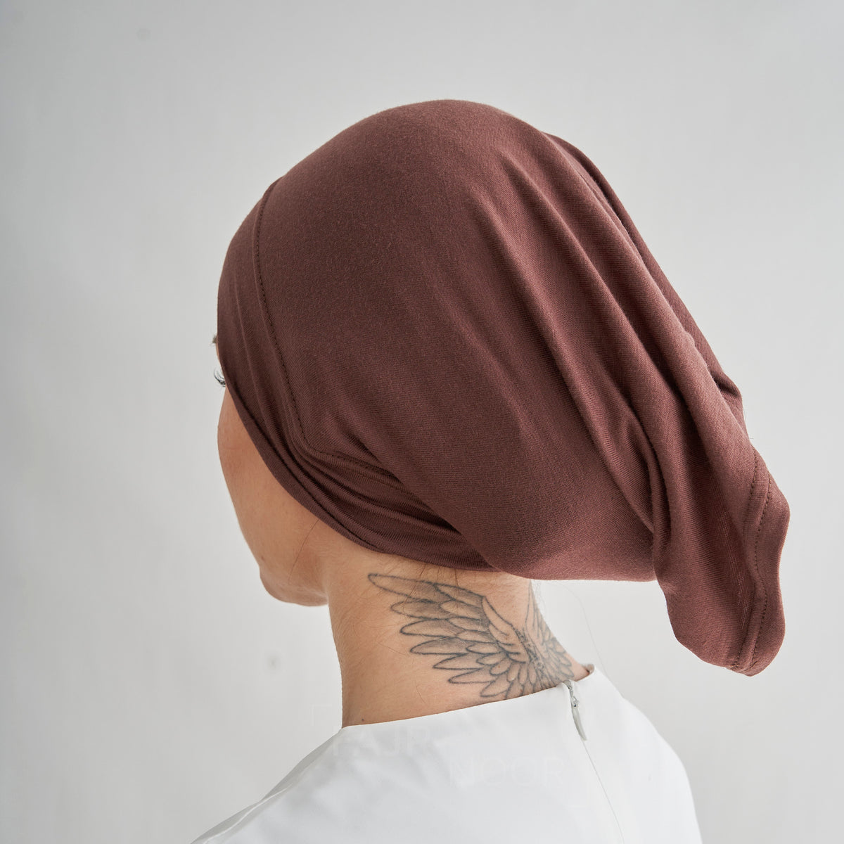 A person shown from the back wearing a draped, slouchy brown fabric head covering or turban-style cap, photographed against a plain light gray background.