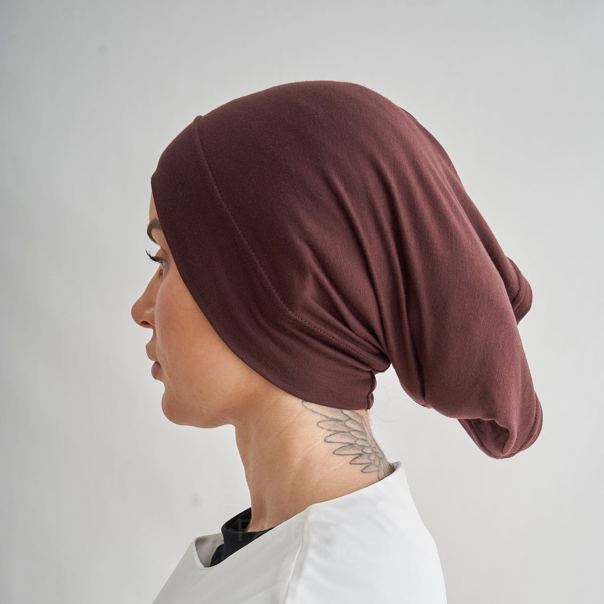 A side profile of a woman wearing a dark brownish-red or burgundy slouchy head covering, also known as a hijab or turban cap, against a plain light gray background.
