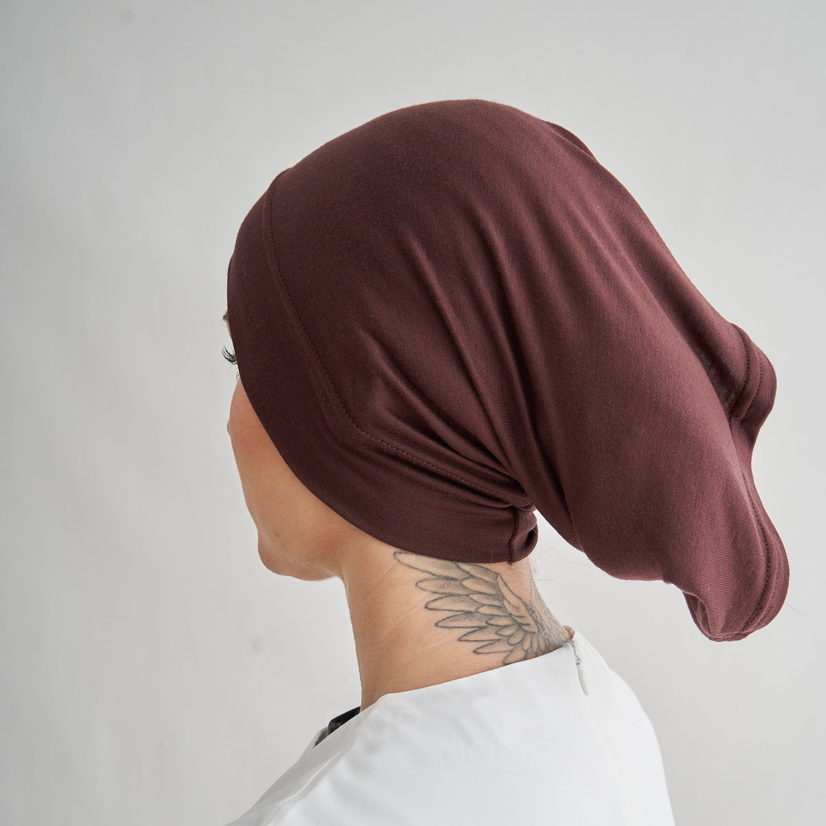 A person shown from the side wearing a dark brown fabric head covering or hijab, photographed against a plain light gray background.