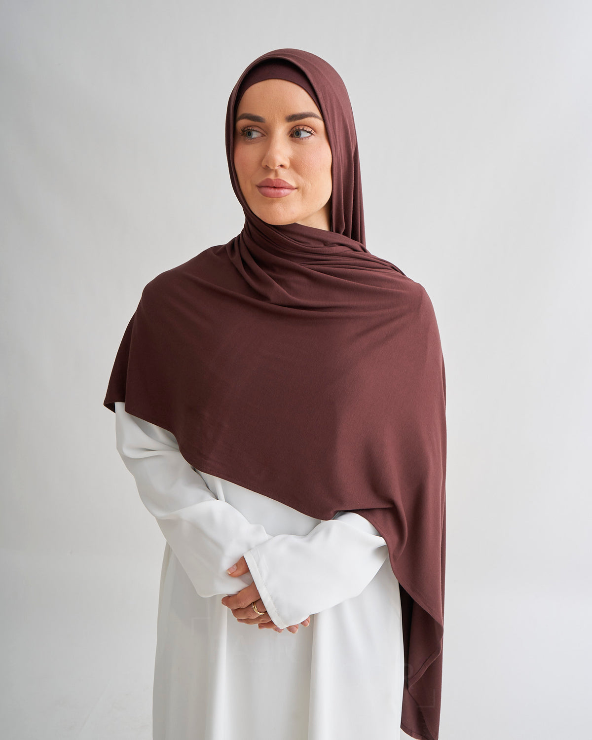 A woman wearing a draped dark brown hijab and a white long-sleeve dress, standing against a light gray studio background.