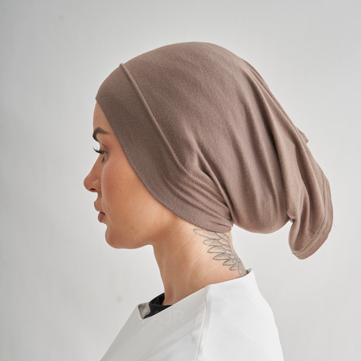 A side-profile view of a woman wearing a soft, draped taupe-brown fabric head covering or hijab against a light gray background.