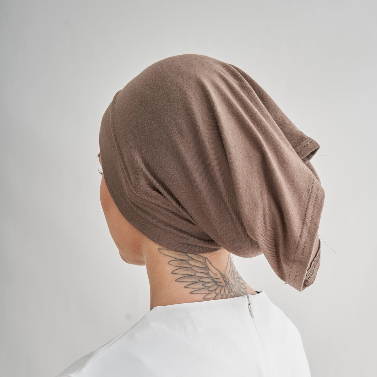 A person shown from the back wearing a soft, draped taupe-brown head covering or hijab, paired with a white top, against a plain light gray background.
