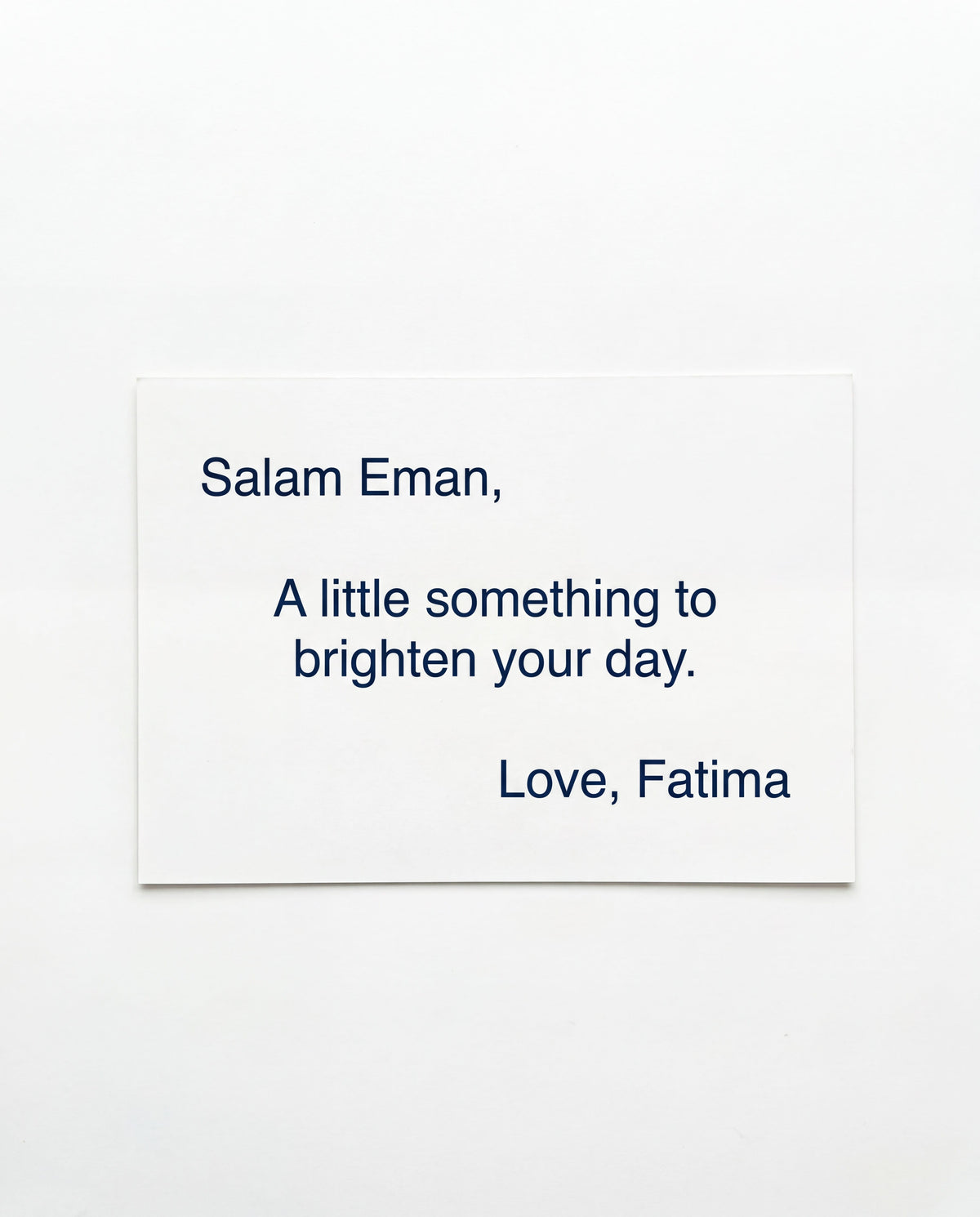 A minimalist white greeting card with navy blue text reading 'Salam Eman, A little something to brighten your day. Love, Fatima' on a clean white background.