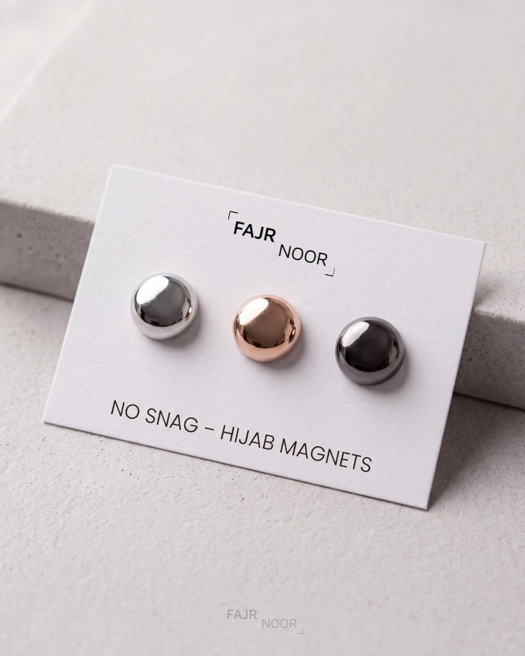 Three small spherical magnetic studs on a white display card labeled FAJR NOOR, with one silver stud, one rose gold stud, and one dark gray/black stud. The card reads 'NO SNAG - HIJAB MAGNETS'.