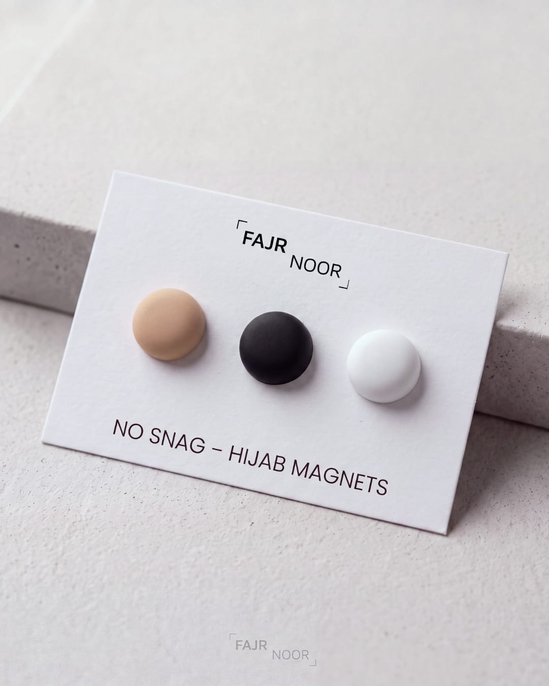 Three small round hijab magnets in beige, black, and white displayed on a white branded card that reads “FAJR NOOR” and “NO SNAG - HIJAB MAGNETS,” photographed on a light neutral surface.
