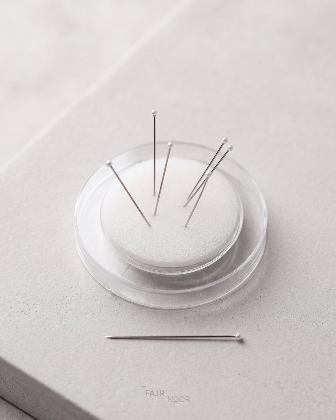 A small clear round container holding several silver sewing pins, with one pin placed outside the container on a light gray surface. The Fair Door logo appears faintly near the bottom.