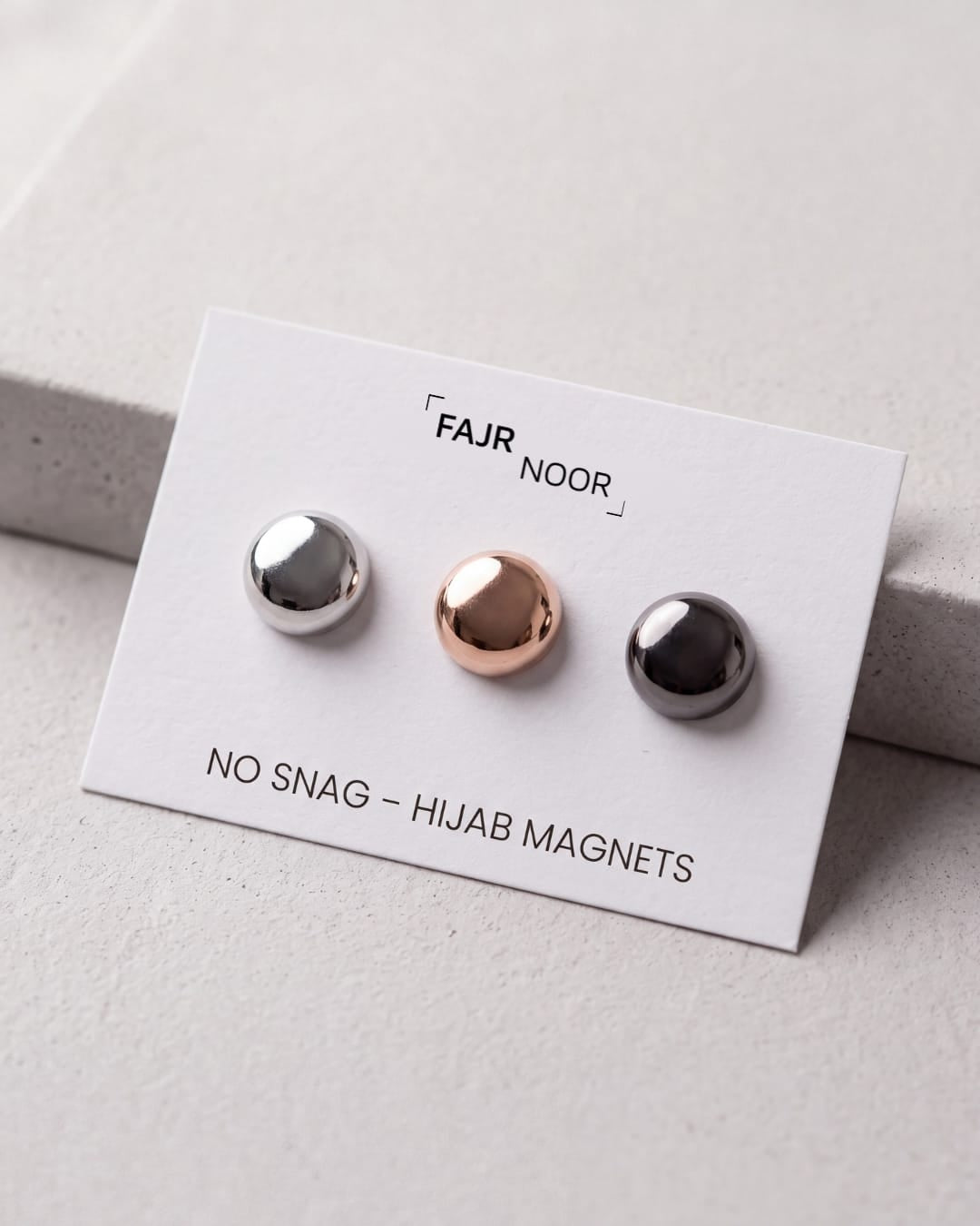 Three small spherical magnetic studs on a white display card labeled FAJR NOOR, with one silver stud, one rose gold stud, and one dark gray/black stud. The card reads 'NO SNAG - HIJAB MAGNETS'.