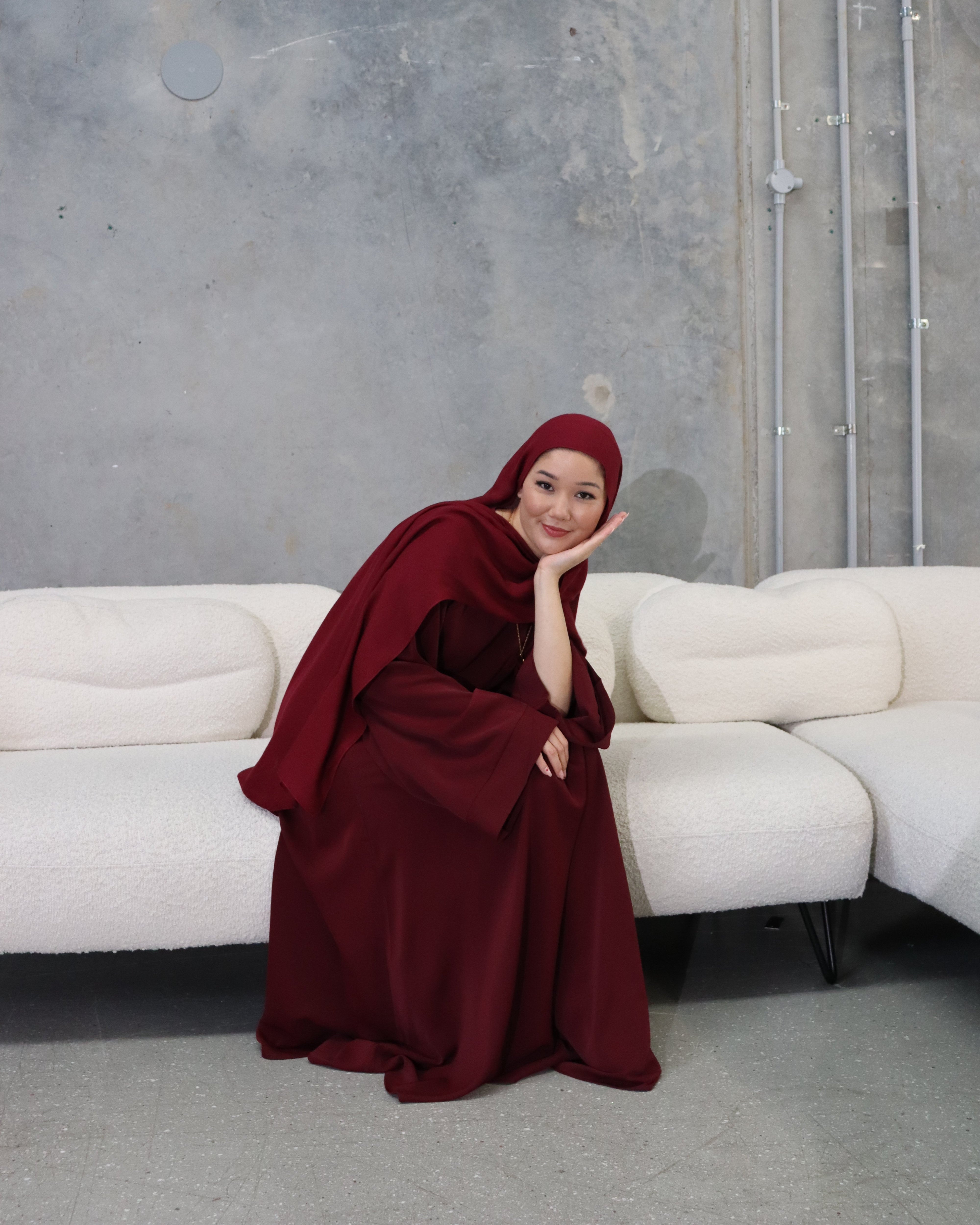 Woman in a burgundy dress and hijab sitting on a white couch against a gray wall.