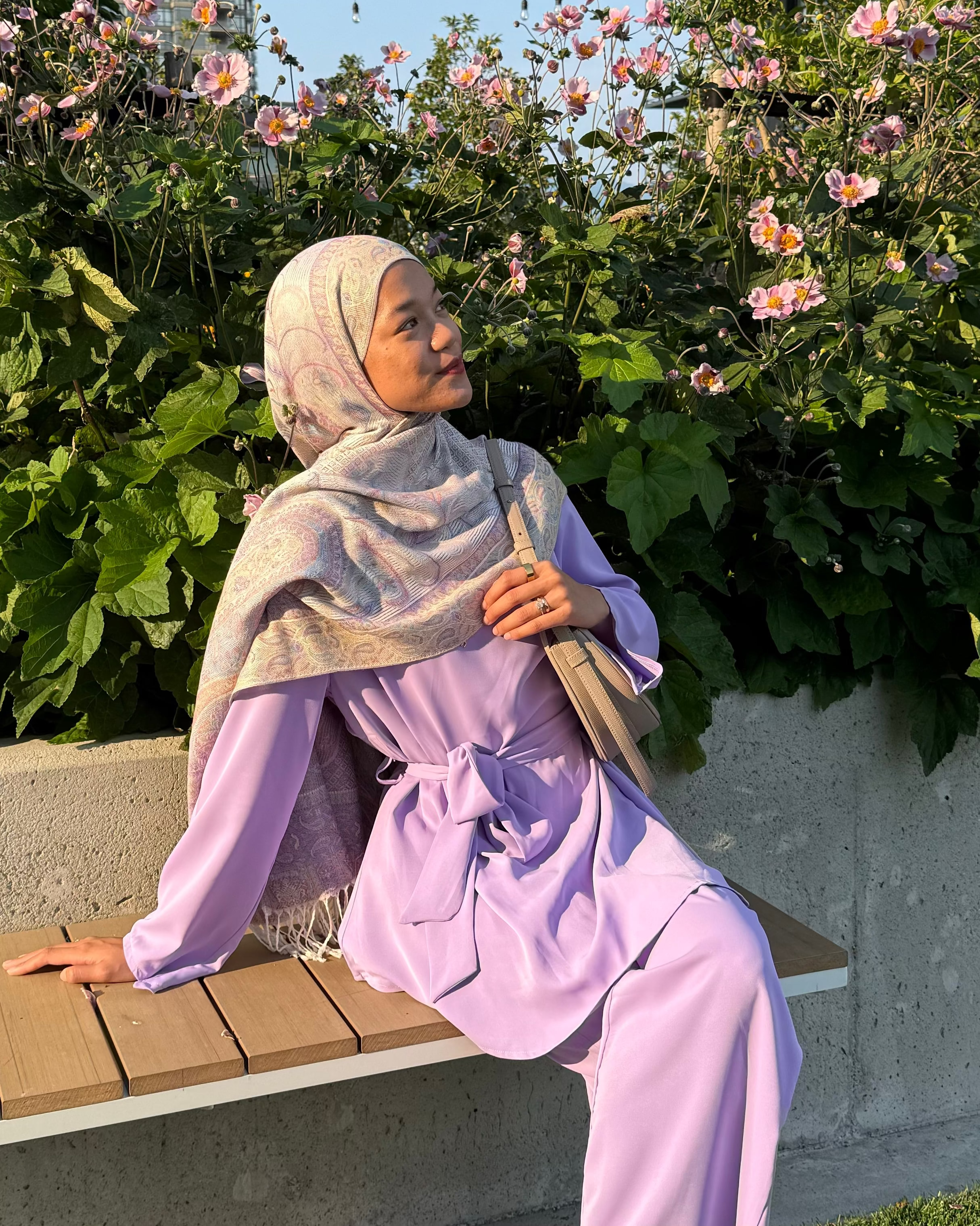 Woman in a lavender outfit sitting on a bench with flowers and greenery in the background