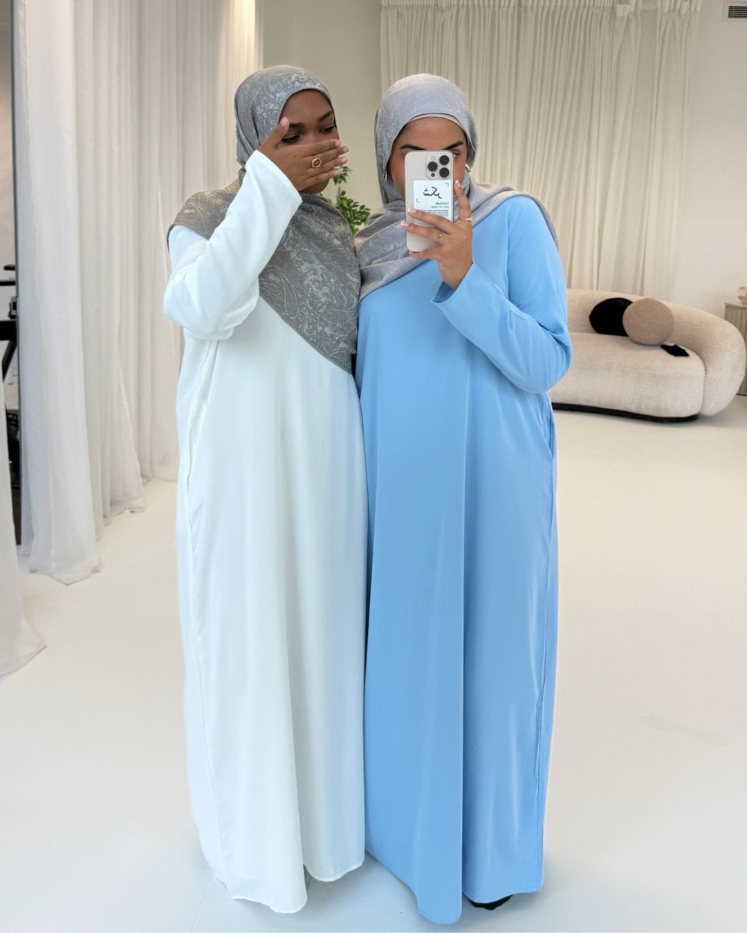 Two women wearing hijabs and long dresses in a room with white curtains.
