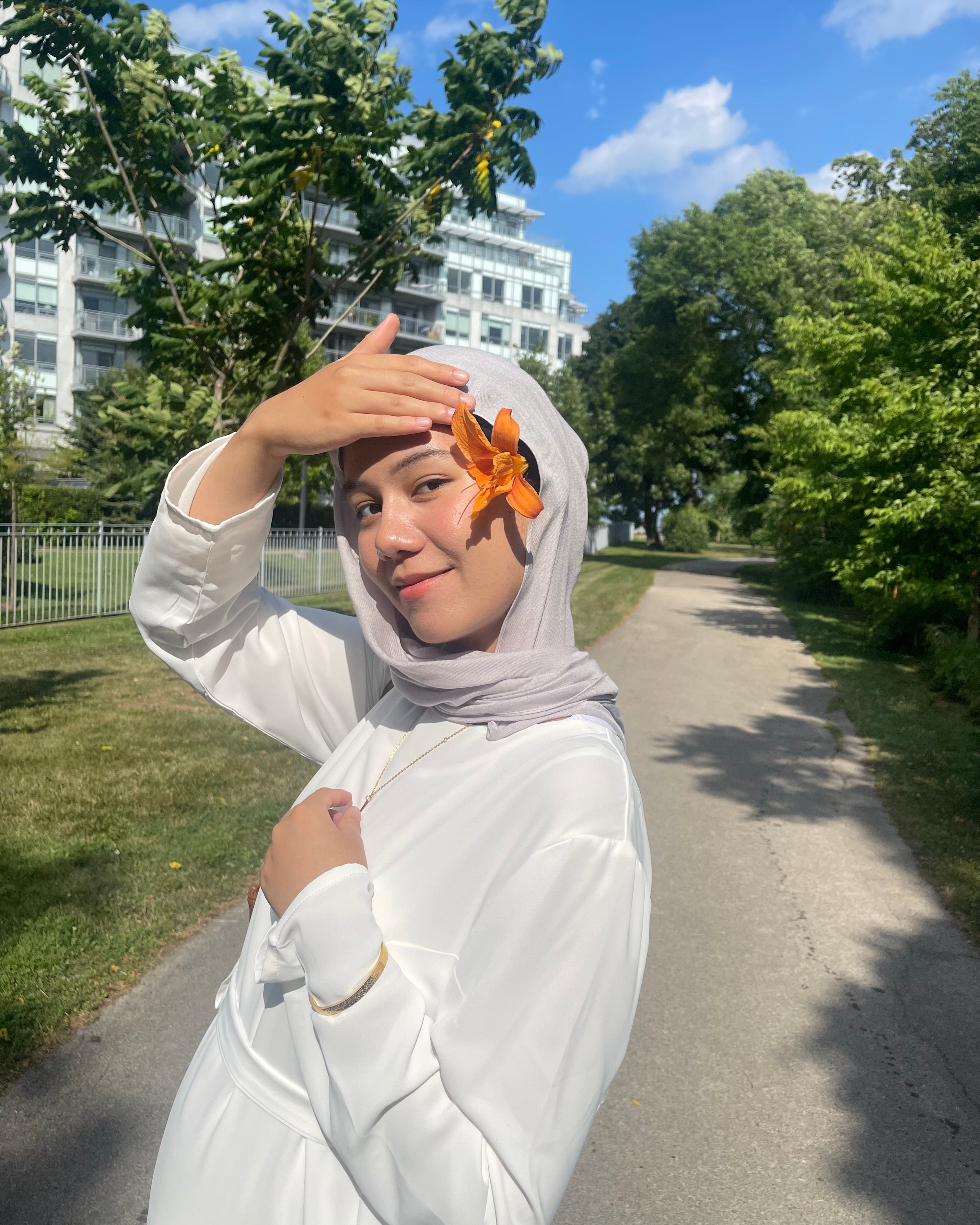 Person wearing a hijab with an orange flower, standing outdoors on a sunny day.