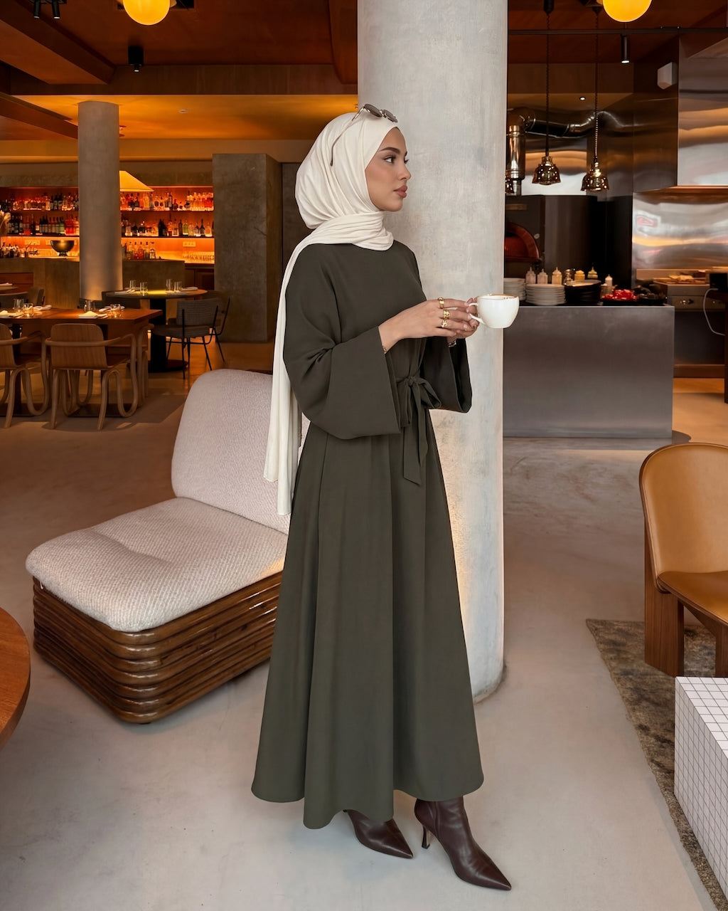 A woman wearing a long-sleeved olive green maxi dress and a white hijab stands in a modern restaurant or cafe setting, holding a white cup.
