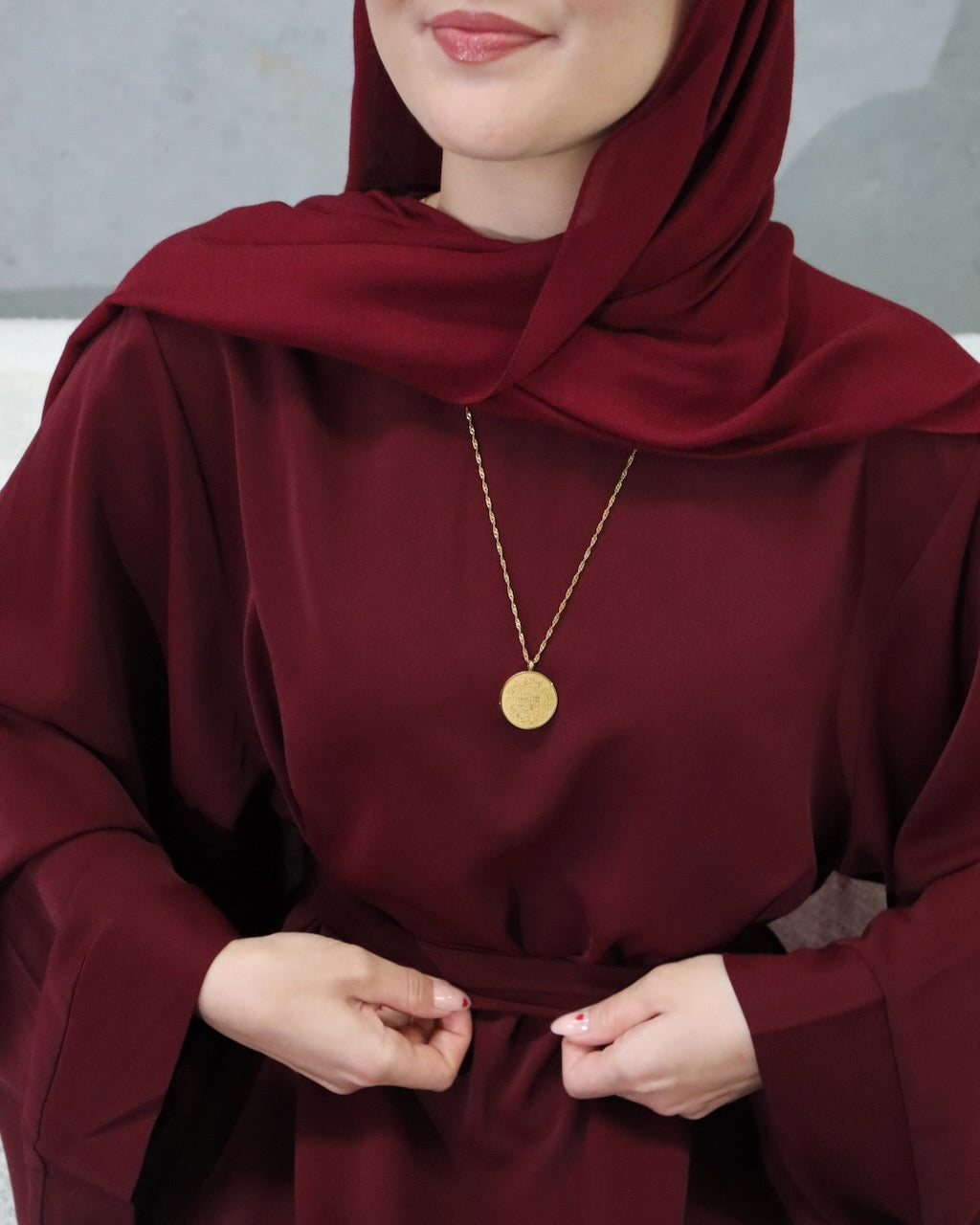 A close-up of a person wearing a deep burgundy abaya with a matching headscarf, accessorized with a delicate gold coin pendant necklace.
