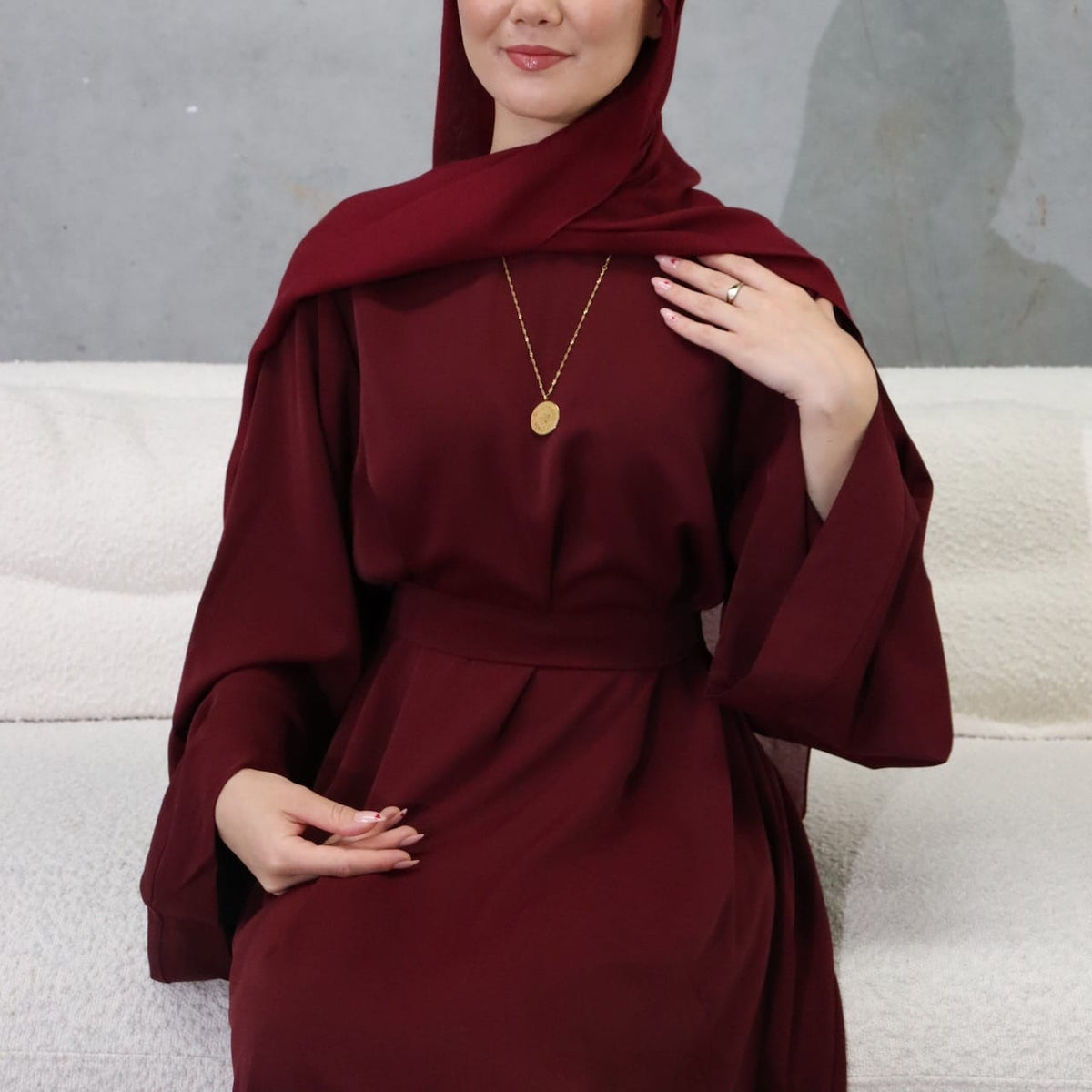 A woman wearing a deep burgundy maroon abaya dress with a matching headscarf and a delicate gold coin necklace, posing on a white textured sofa against a gray wall.