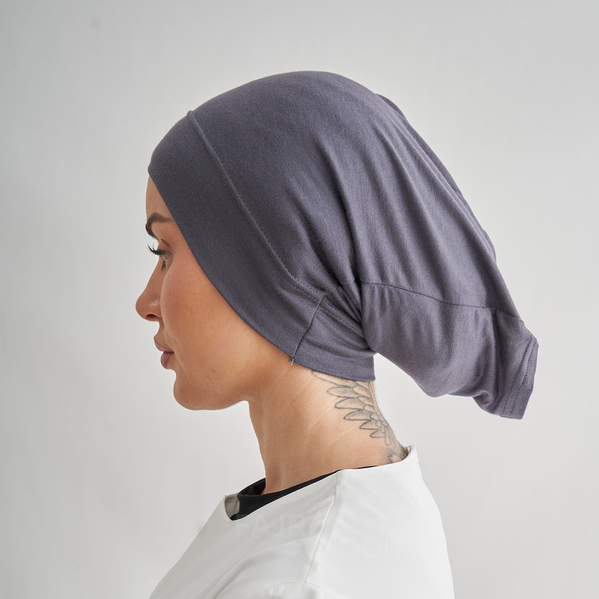 A side profile of a woman wearing a dark slate gray fabric head covering or hijab, paired with a white top, against a light gray studio background.
