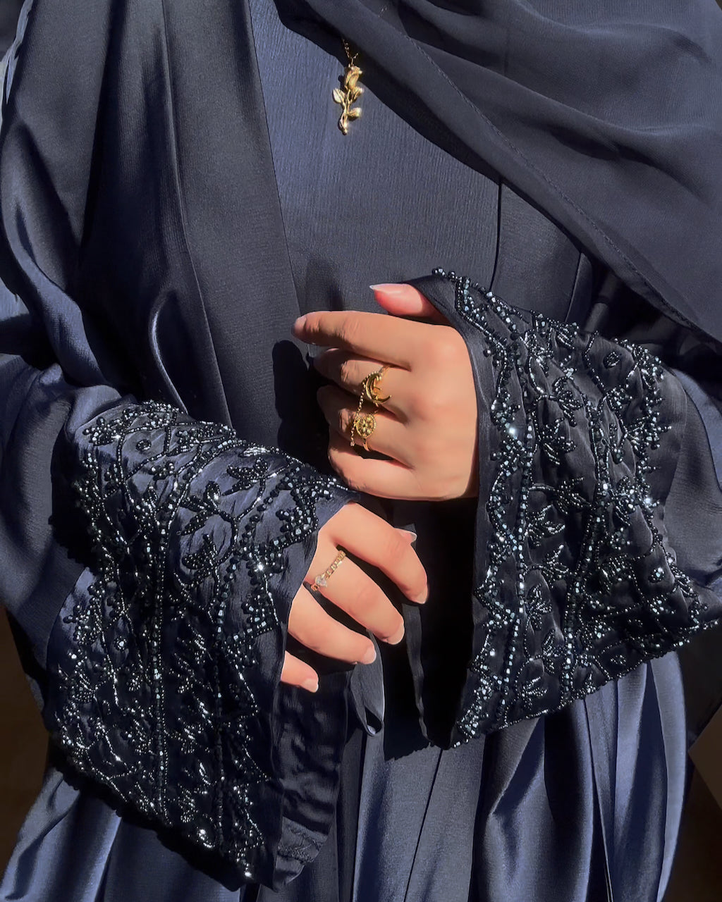 A close-up of a person wearing a dark navy blue abaya with heavily embellished beaded and sequined bell sleeves, a matching hijab, and gold rings.
